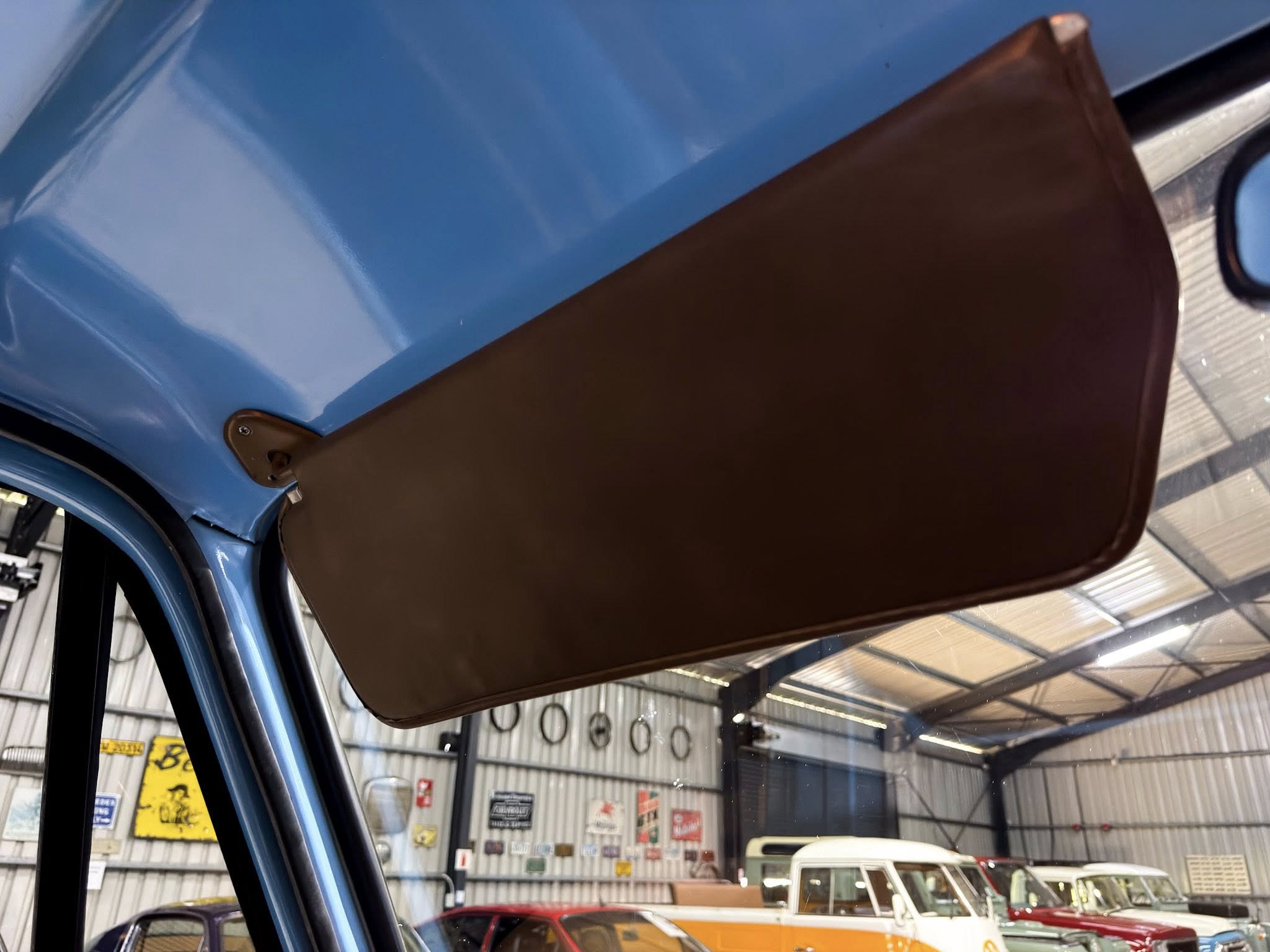 Interior of a blue car with a brown sun visor extended, inside a workshop with vintage vehicles visible in the background.