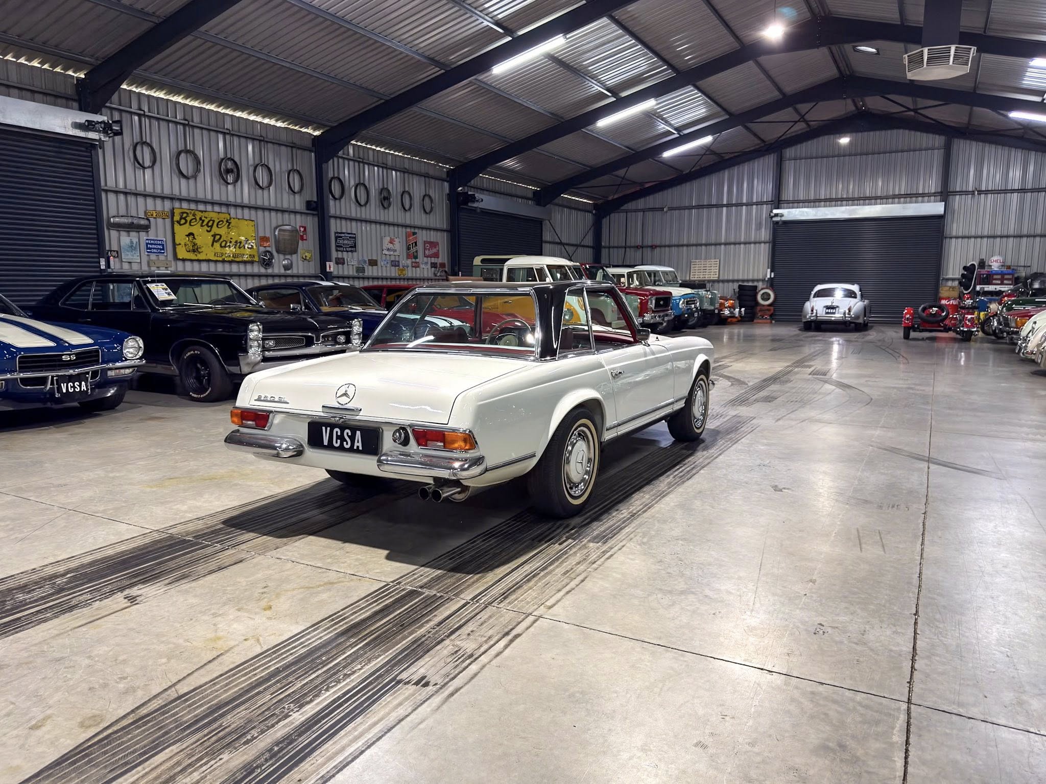 Indoor classic car showroom with a white Mercedes convertible in the foreground and rows of vintage cars along metal walls and signs in the background.