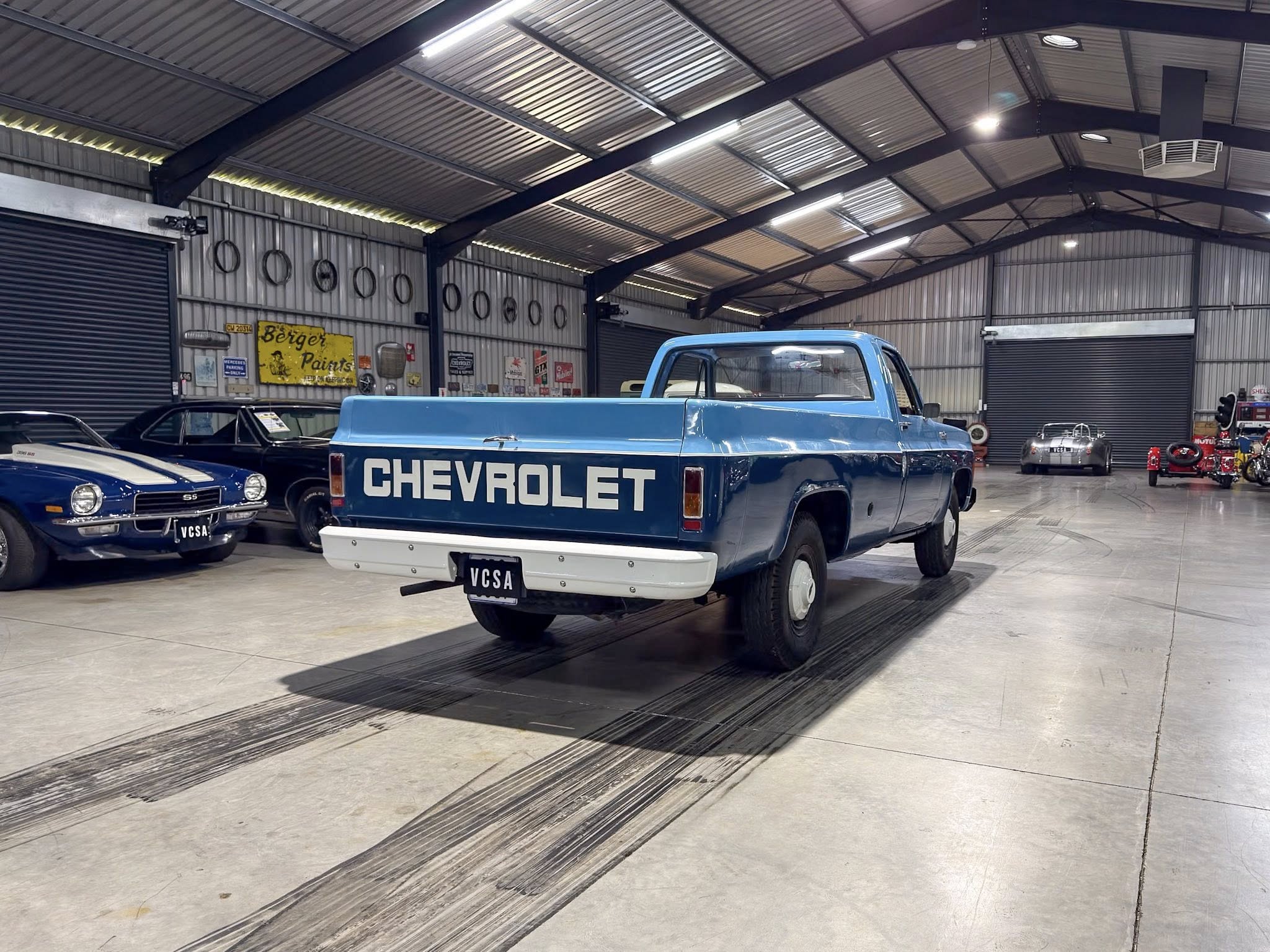 Blue Chevrolet pickup parked in a large indoor showroom with other classic cars and vintage signs on the walls.
