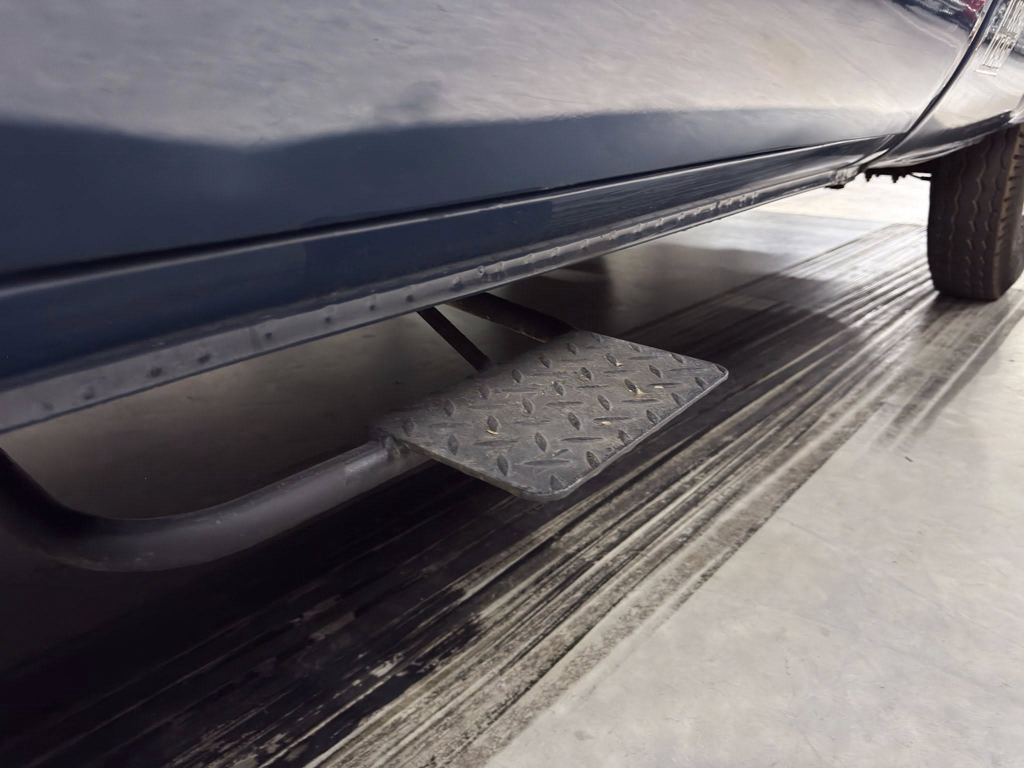 Close-up of a blue vehicle's lower side with a diamond-plate step and a tire on a concrete loading dock area.