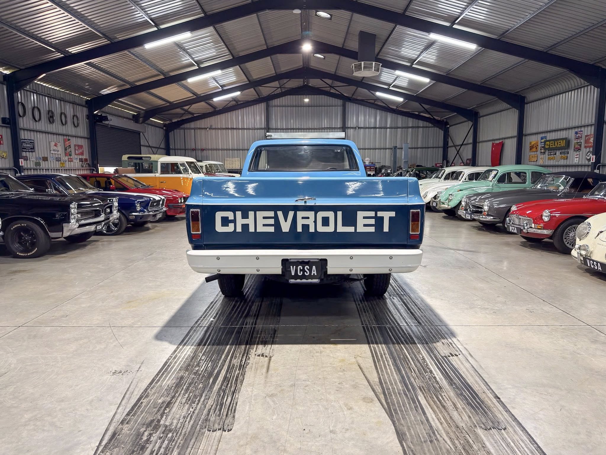 Blue Chevrolet pickup in a spacious showroom surrounded by vintage cars on both sides of the room.