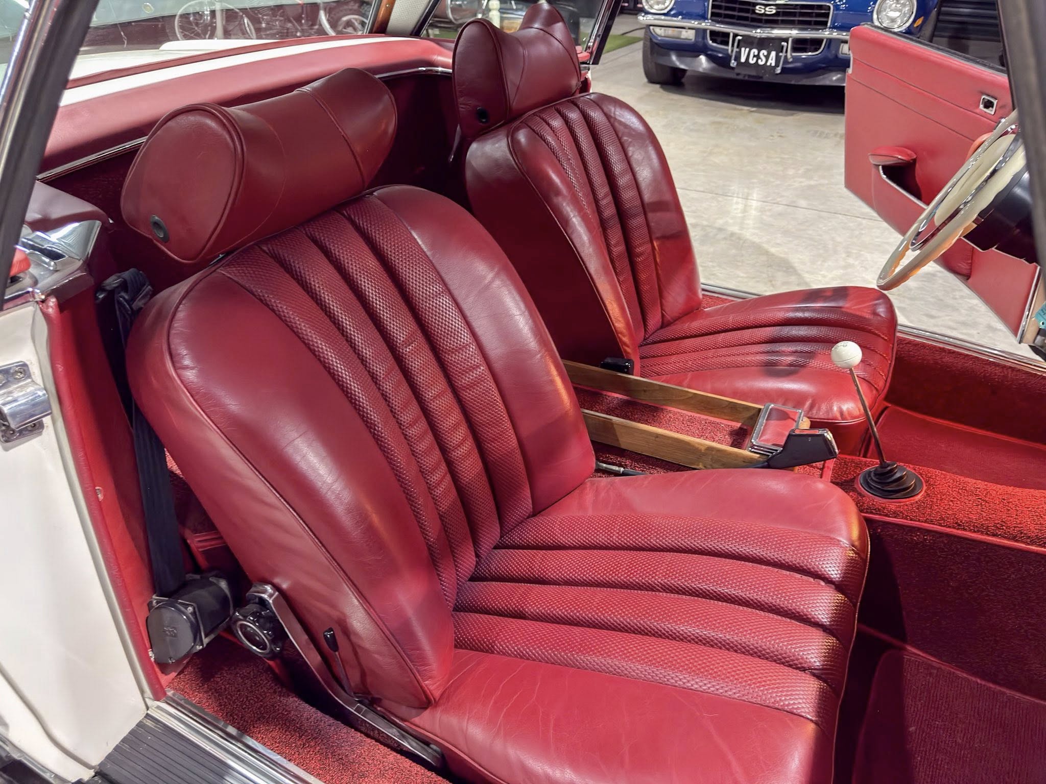 Inside a vintage red car showing red leather seats, a manual gear shifter, and an open door edge close to the frame.