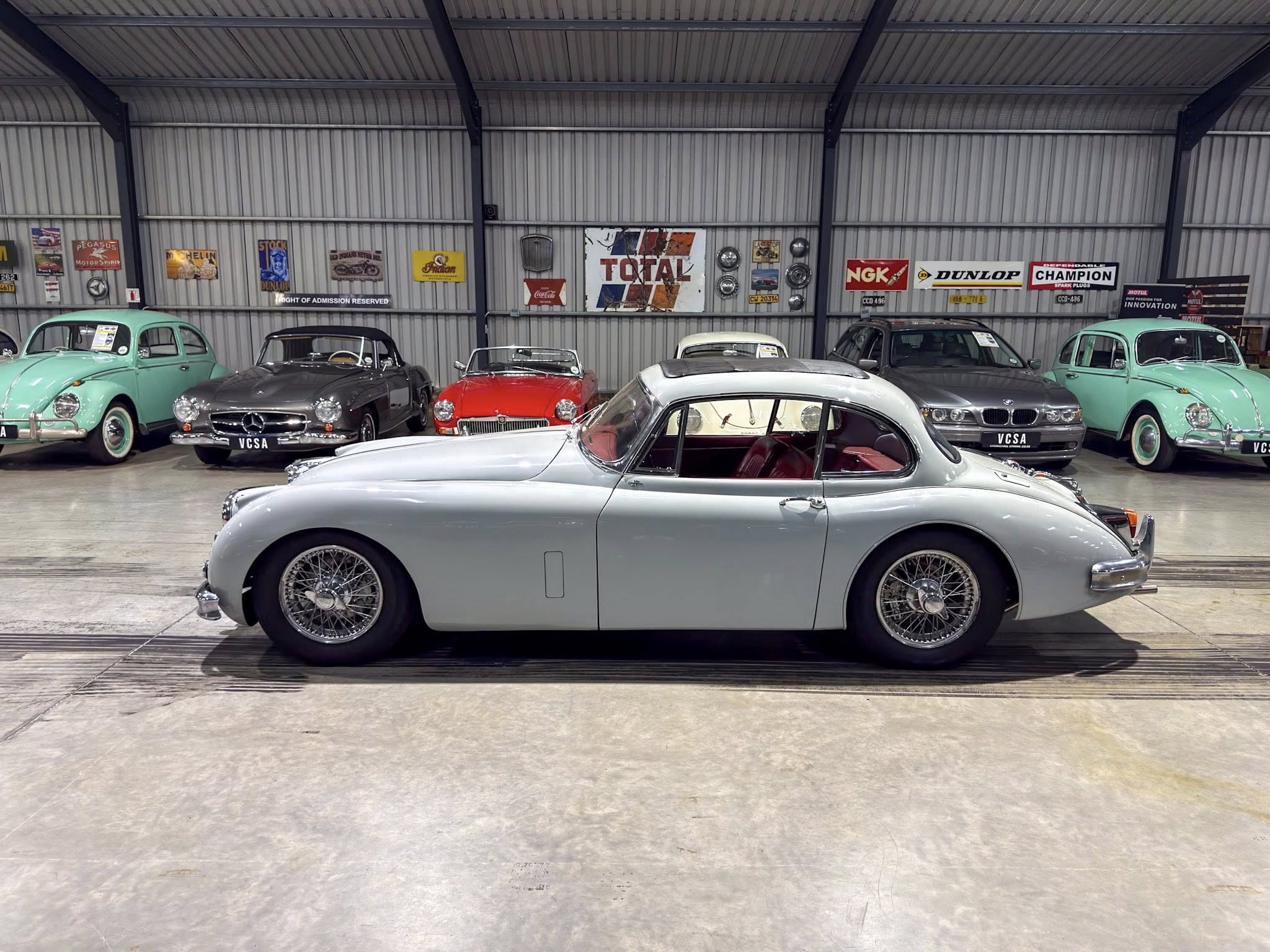 Indoor garage with a row of vintage cars; foreground features a large silver classic car, with mint green cars and more in the background against corrugated metal walls with retro signs.