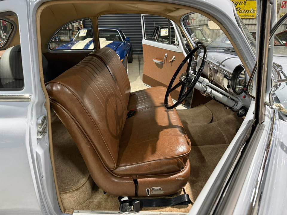 Open-door view of a classic gray car interior with a brown leather bench seat, black steering wheel, and chrome dashboard in a showroom.