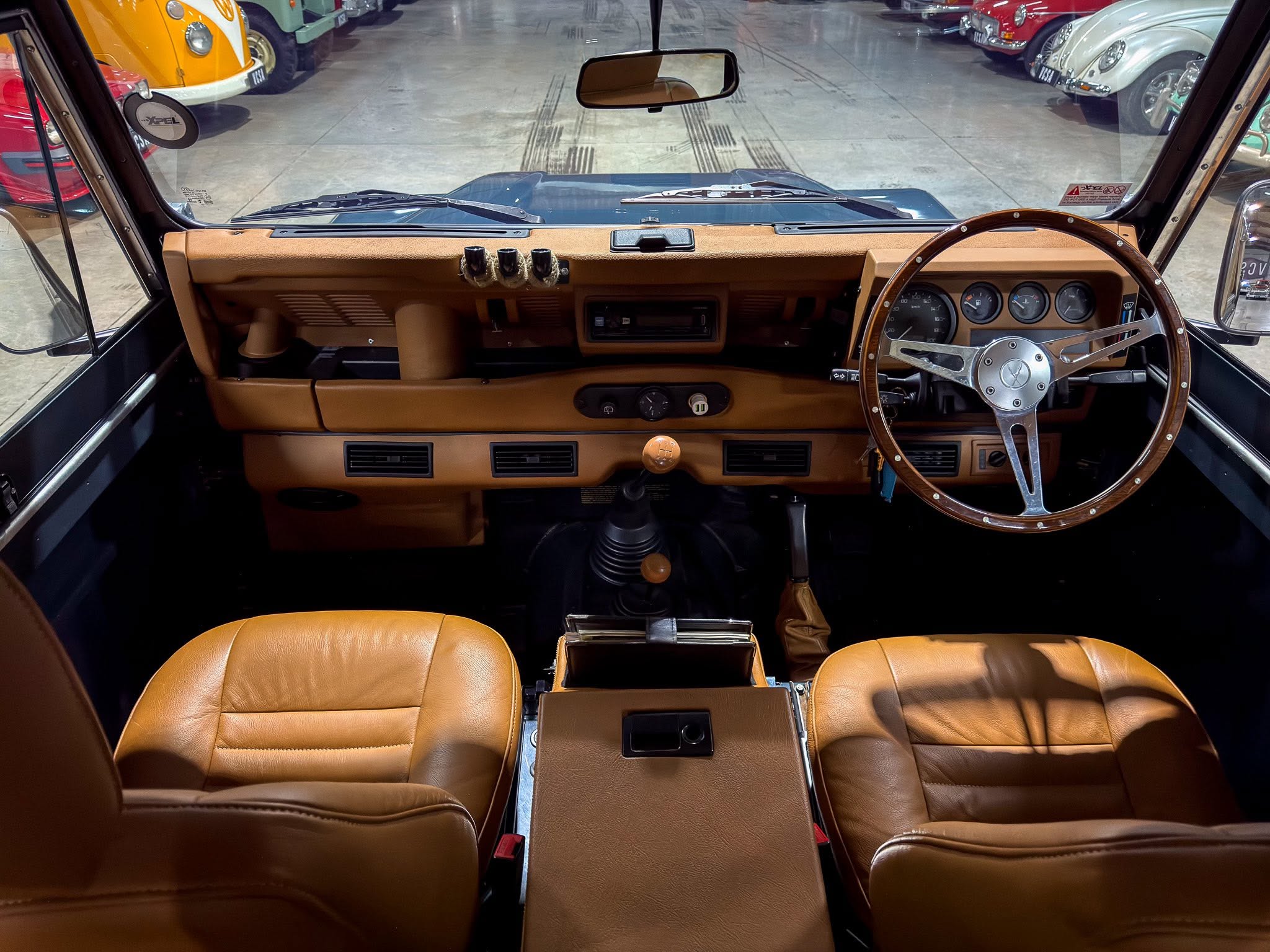 Front interior from the driver’s seat of a vintage car with tan leather seats, a wood-rimmed steering wheel, and gauges; showroom with classic cars visible through the windshield.