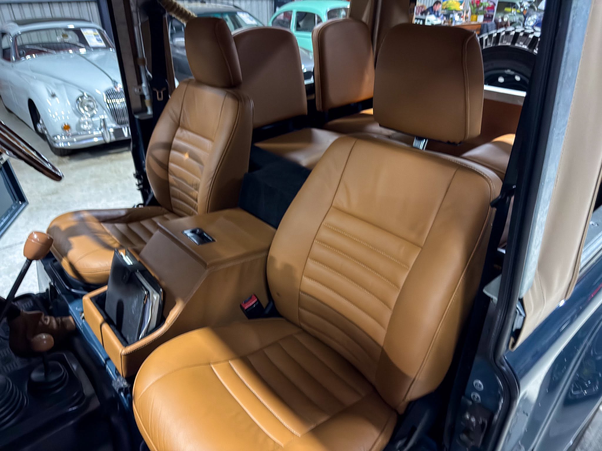 Tan leather car interior with front passenger seat and headrest, viewed from the open door in a vintage showroom setting with classic cars nearby.