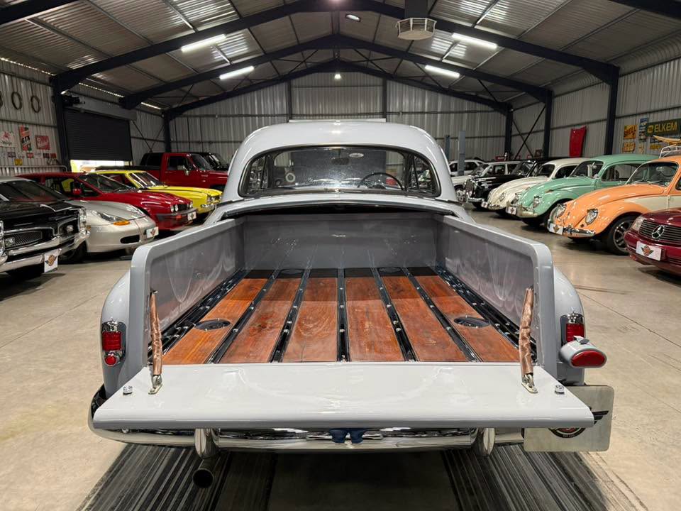 Rear view of a light gray vintage pickup with an empty wood-plank truck bed inside a classic car showroom filled with colorful vintage cars on either side, under industrial lighting.
