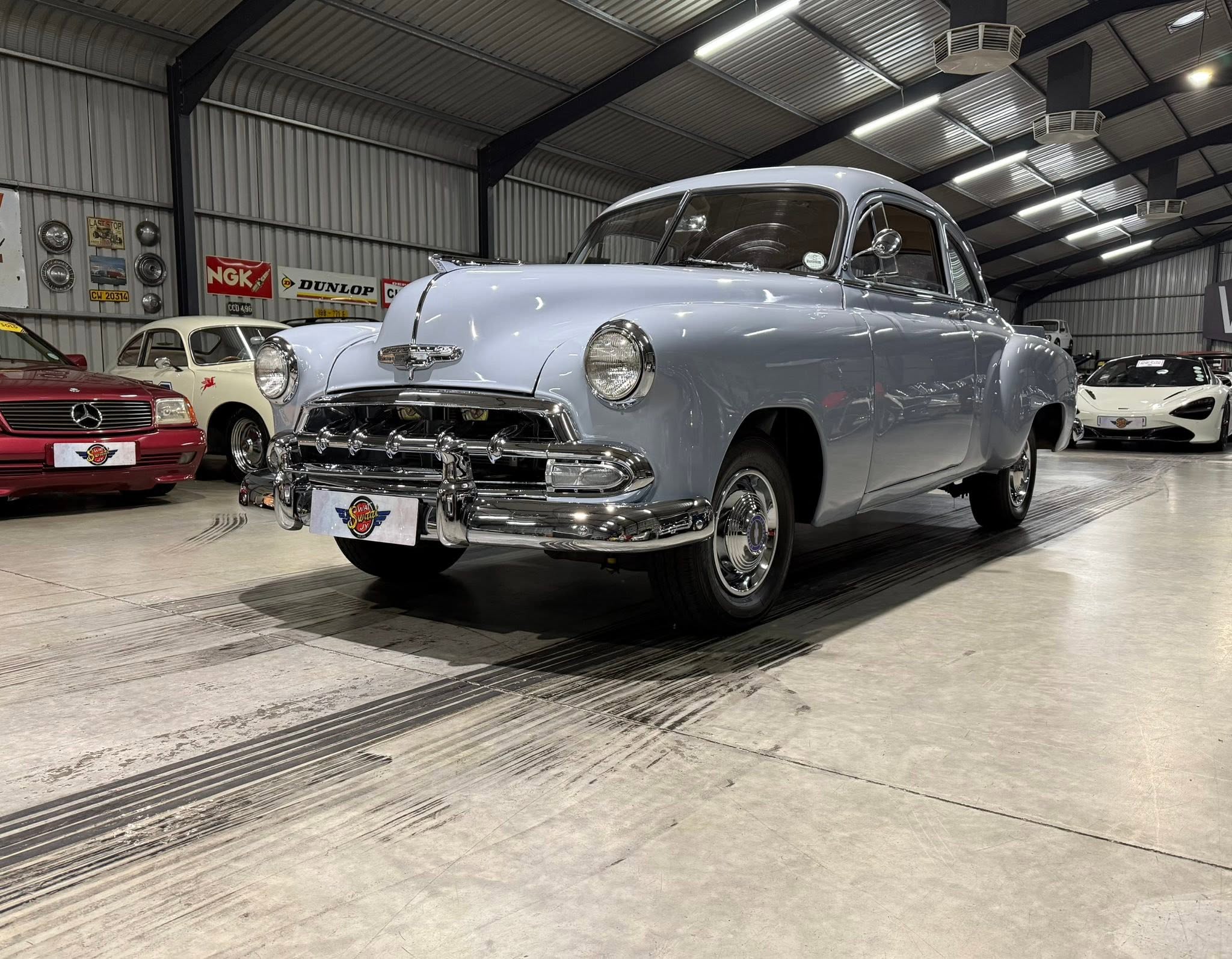 Inside a metal-arched showroom filled with classic cars, featuring a large light blue vintage car in the foreground and other classic vehicles along the wall behind it