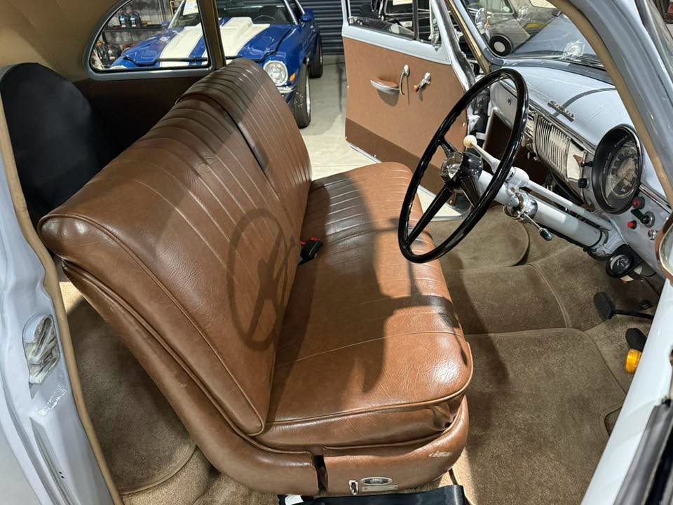 Interior of a vintage car with brown leather bench seat, large steering wheel, and classic dashboard in a showroom setting. Other classic cars are visible outside the car door.