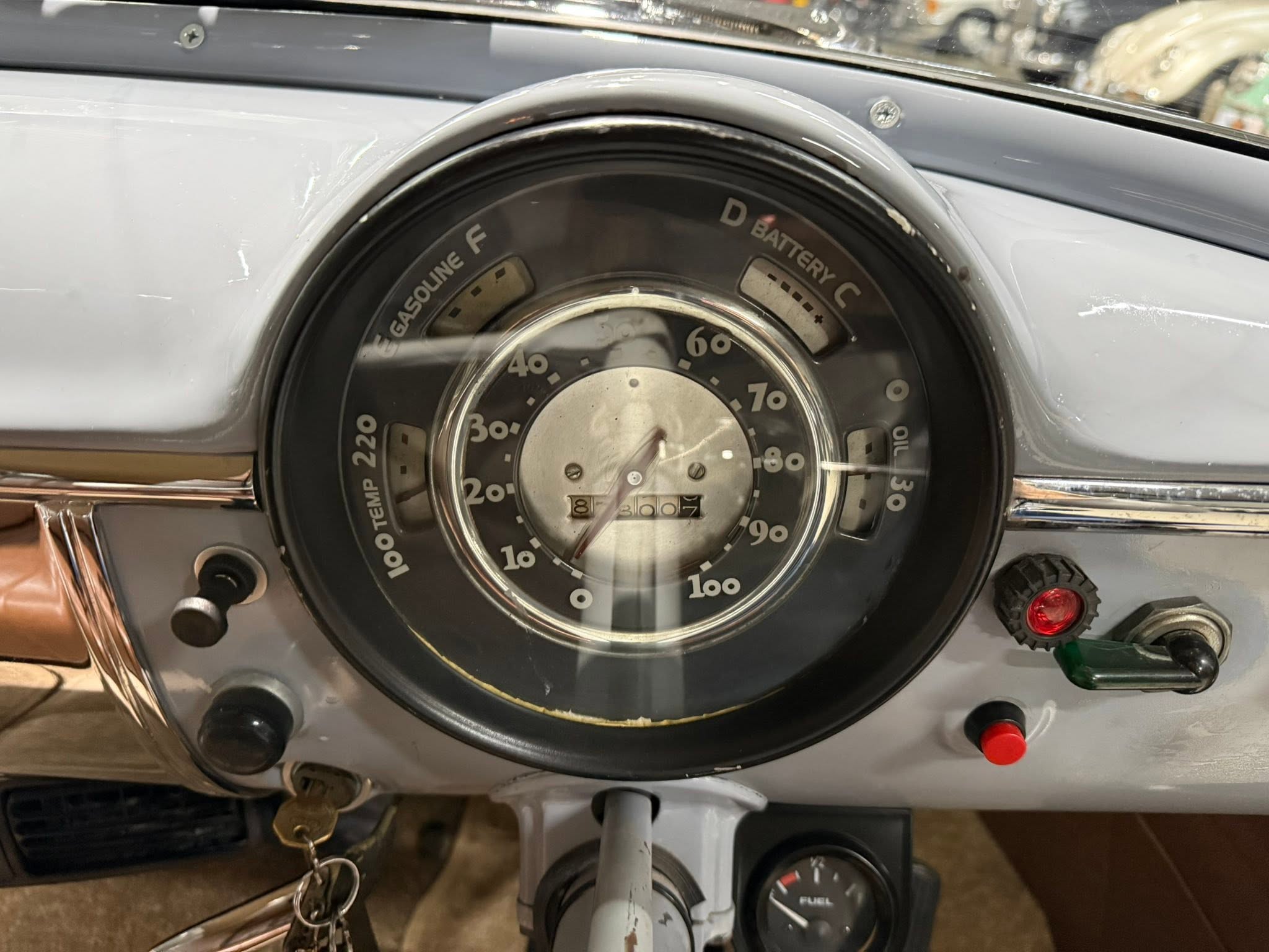 Close-up of a vintage car dashboard: large round speedometer with fuel gauge and ignition area, keys hanging from the ignition switch, and surrounding gray paneling.