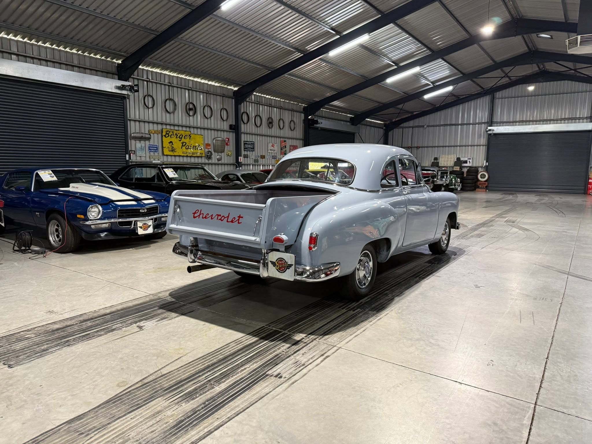 Inside a metal-structured garage/showroom with several vintage cars, including a light blue Chevrolet pickup in the foreground and a blue classic car on the left.