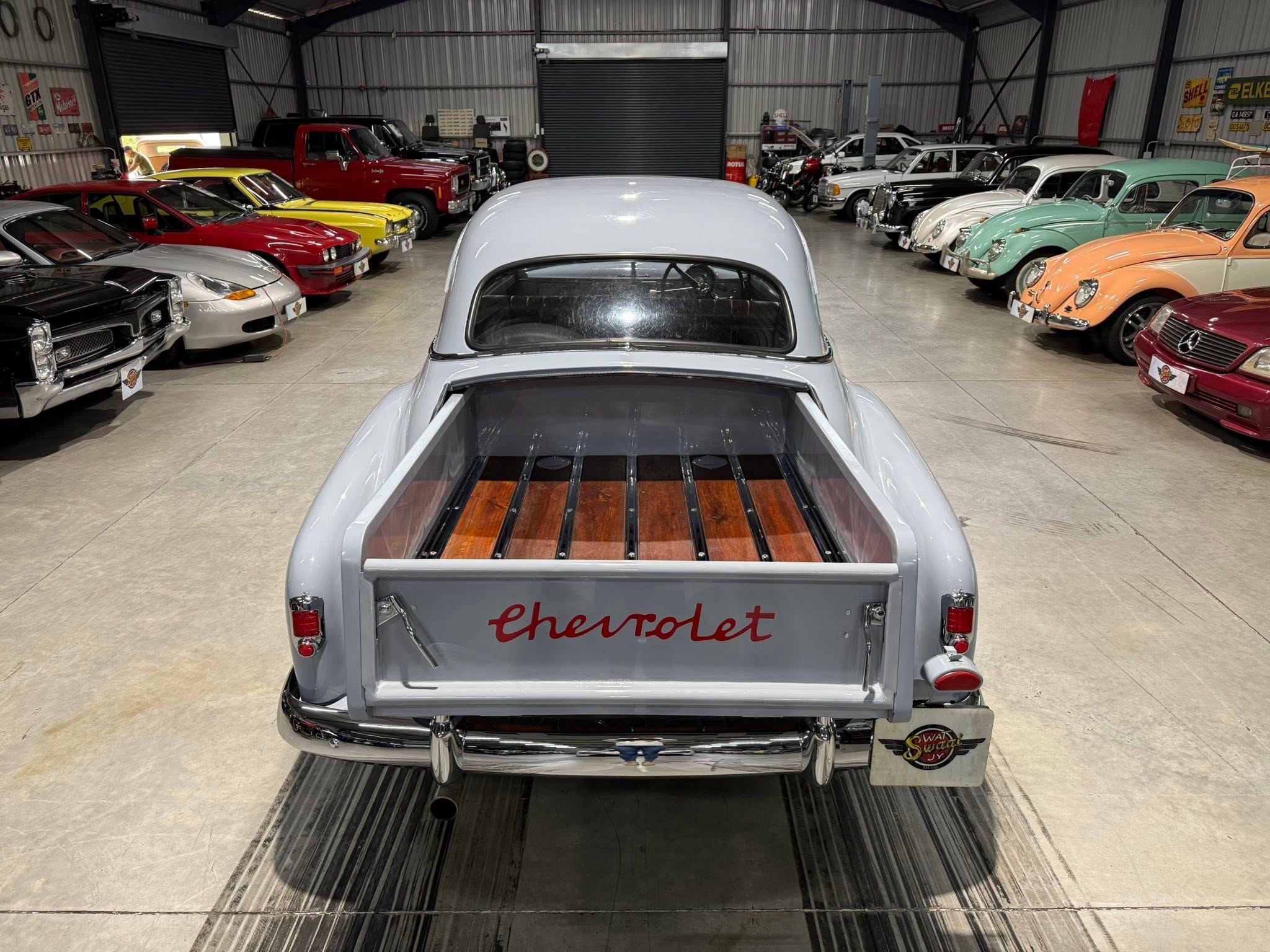 View from the back of a light blue Chevrolet pickup in a showroom filled with rows of vintage cars in multiple colors, indoors.