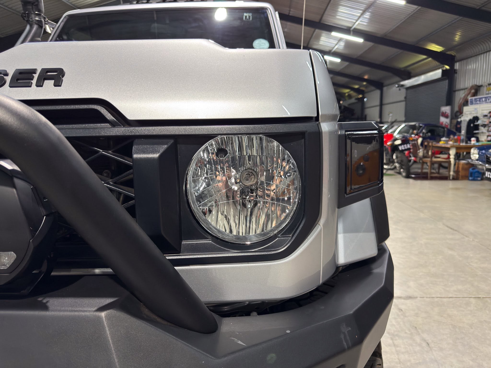 Close-up of the front left corner of a silver Jeep Wrangler, showing the round headlight and bumper in a showroom.