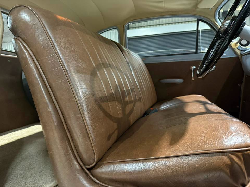Front passenger area of a vintage car with brown leather bench seat and steering wheel visible on the right side in daylight.