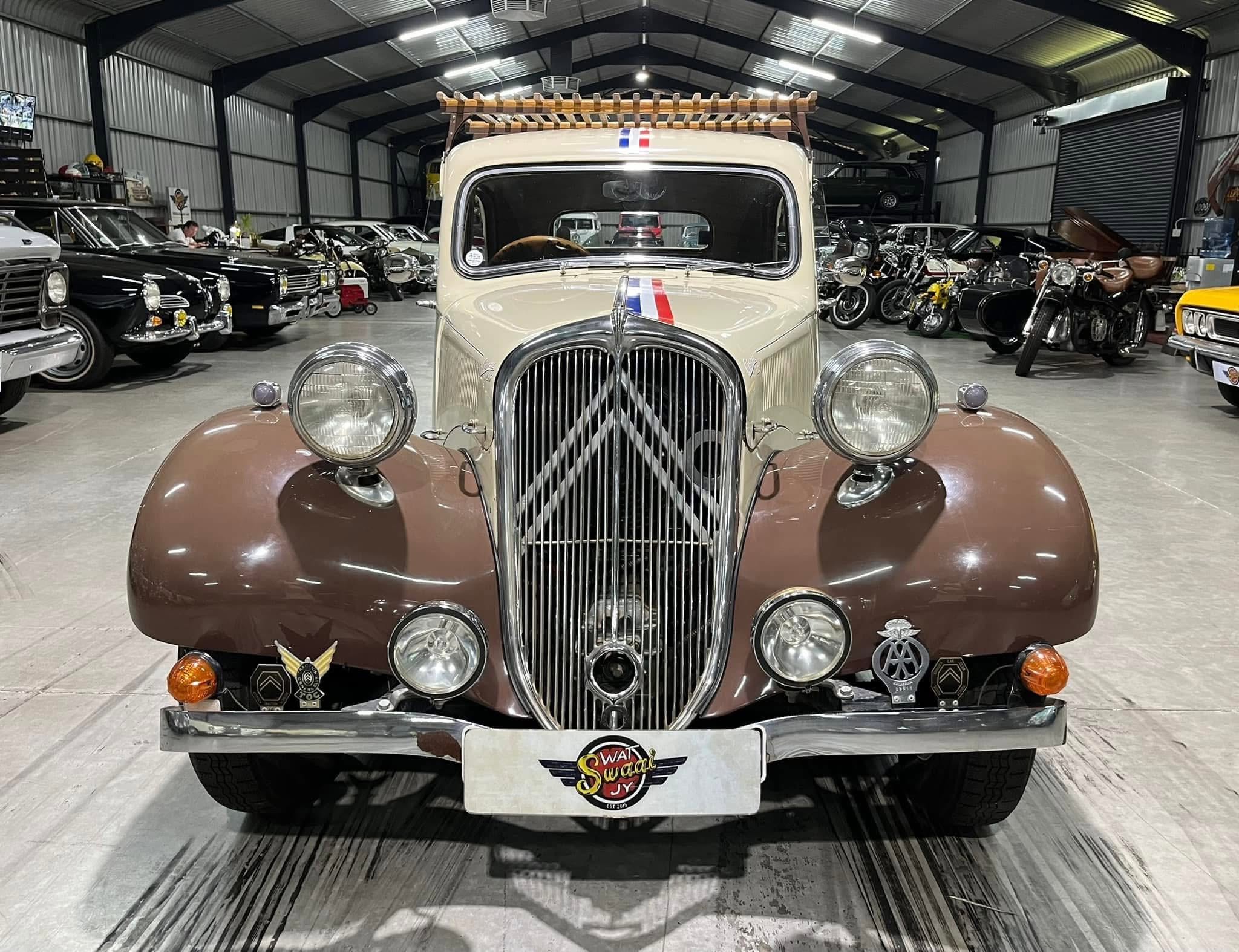 Front view of a cream-colored vintage car with a chrome grille, inside a showroom filled with classic vehicles.