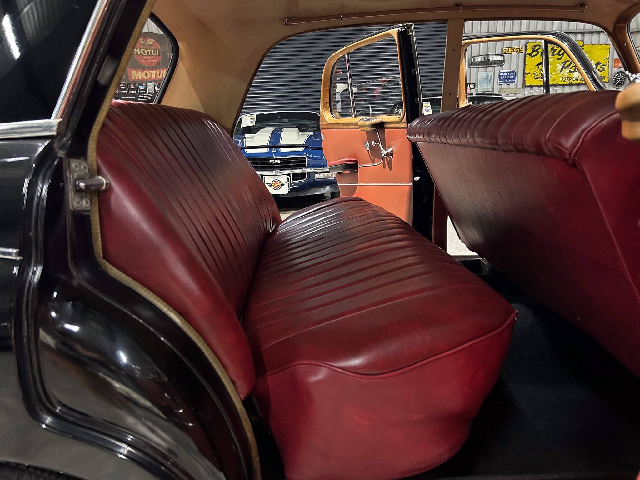 Interior of a vintage car with red leather seats and wood trim, seen from the backseat toward a blue car outside in a showroom.