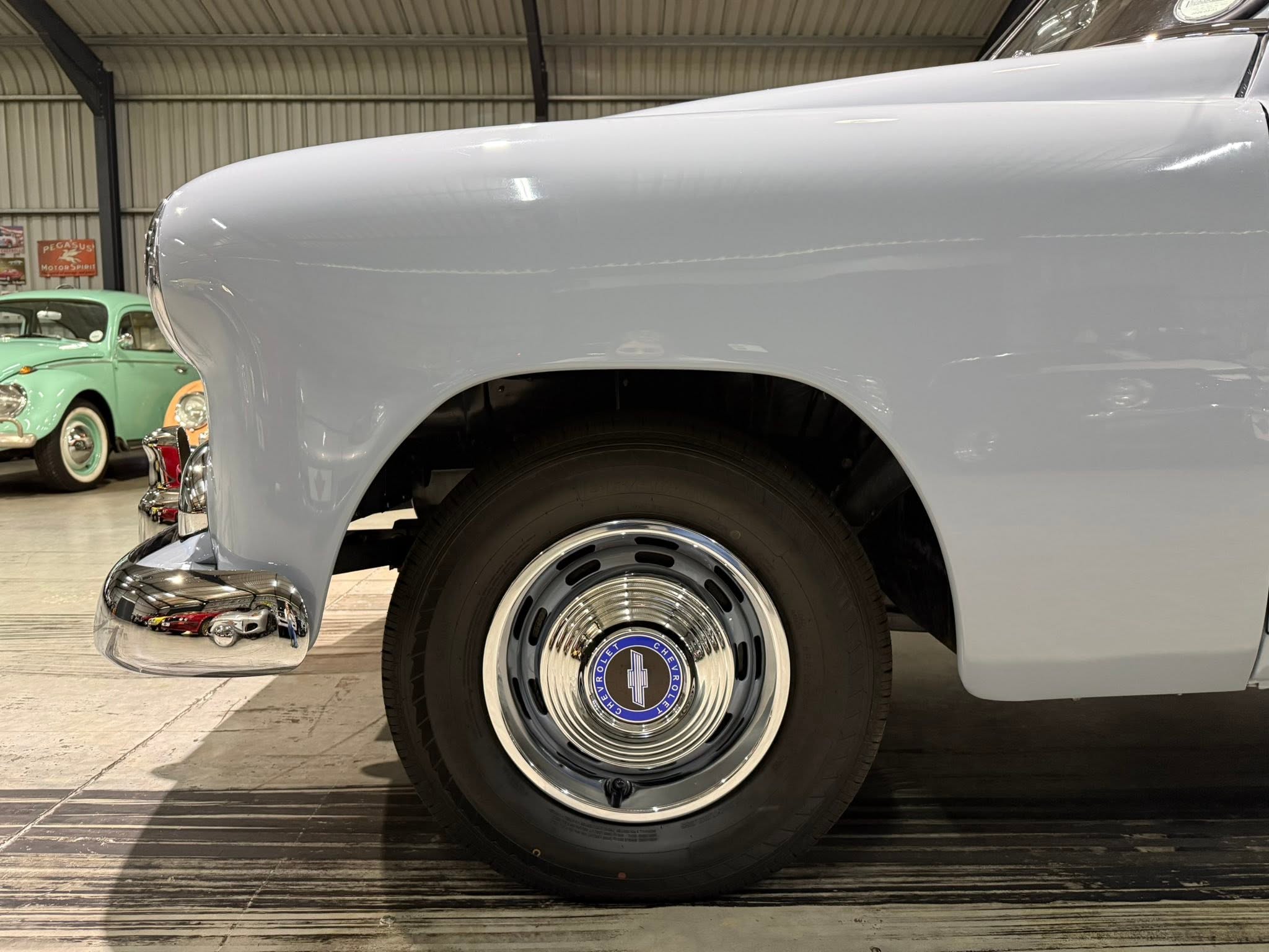 Close-up of a vintage car's front fender and wheel in a showroom, light blue paint and polished hubcap reflecting other classic cars in the background.