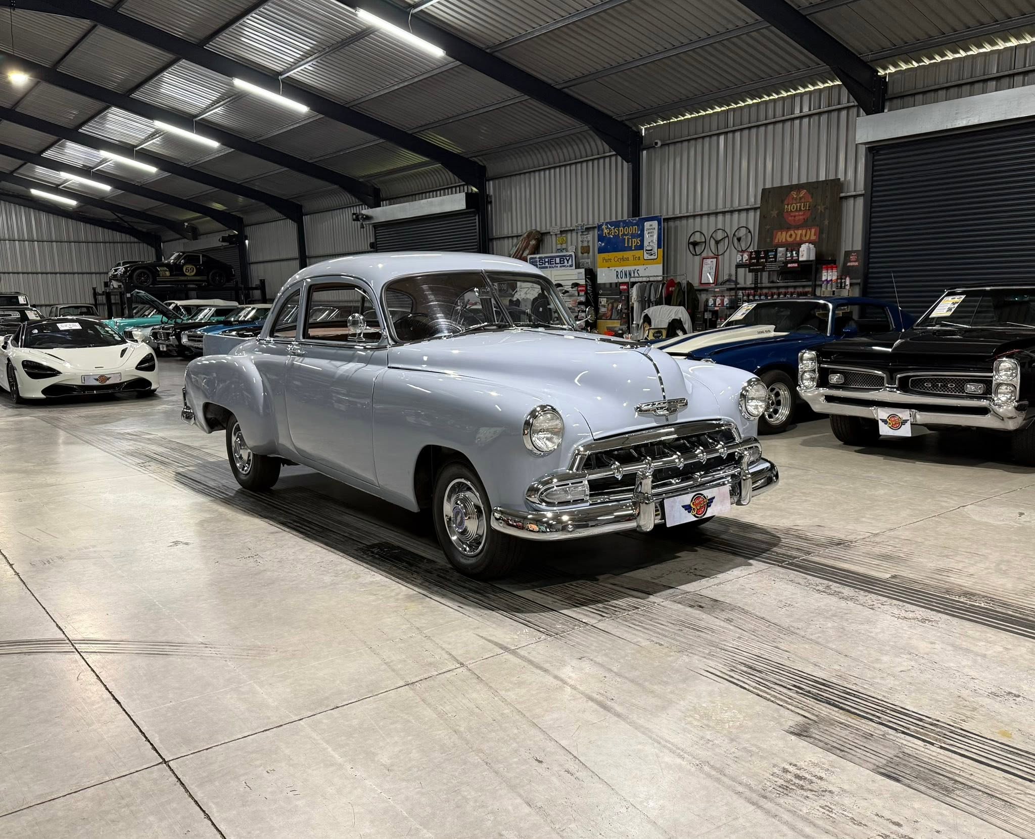 Inside a large metal-structured showroom filled with vintage cars; a light blue classic car dominates the foreground with more classics in the background.