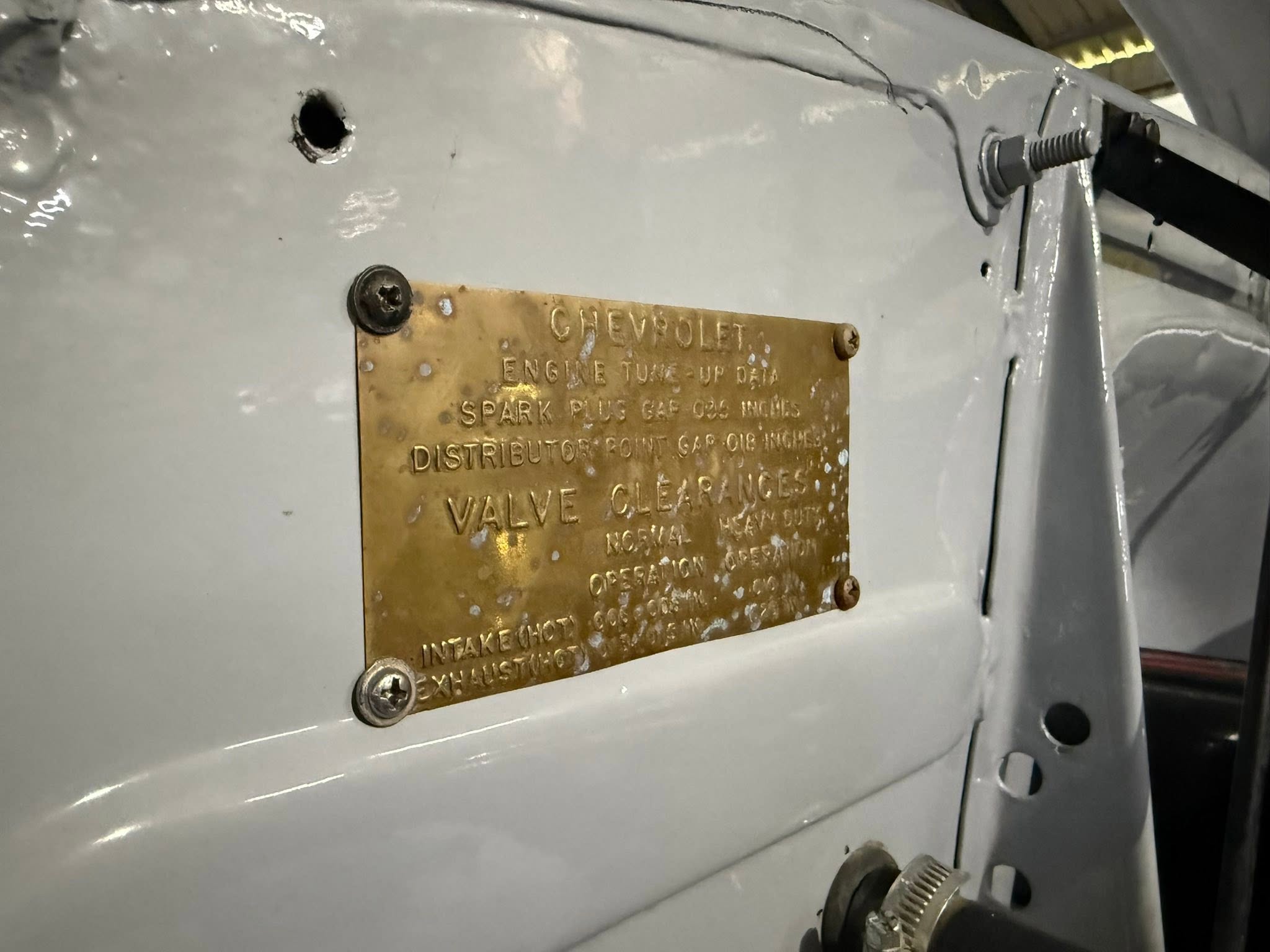 Bronze plaque on a gray metal surface reading 'CHEVROLET ENGINE TUNE-UP DATA SPARK PLUG GAP INCHES' held by screws.