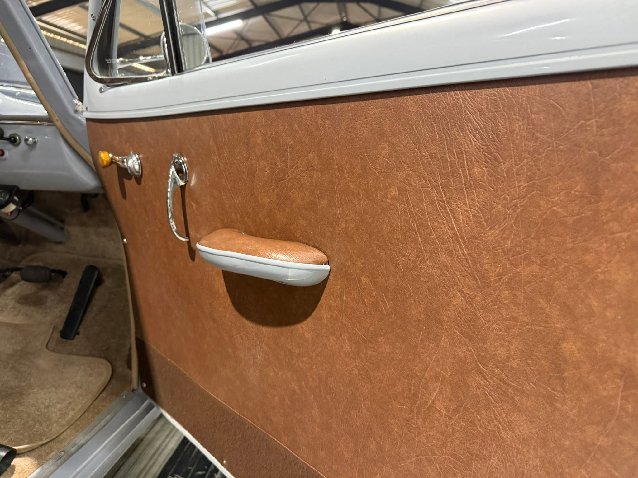 Close-up of a vintage car door: brown textured leather panel, chrome window crank, orange knob, and door handle.