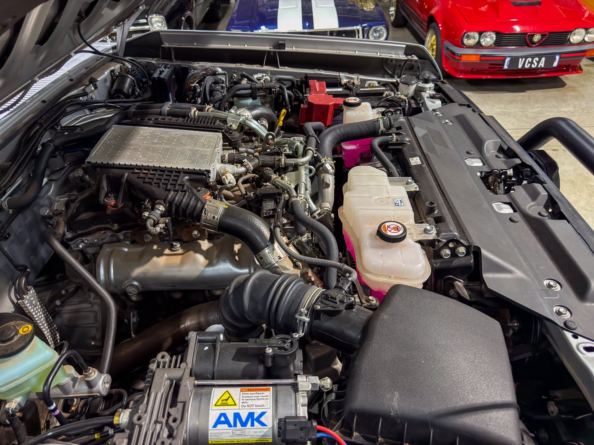 Open hood view of a car's engine bay with exposed hoses, pumps, and a pink coolant reservoir; a labeled AMK battery is visible in the foreground.