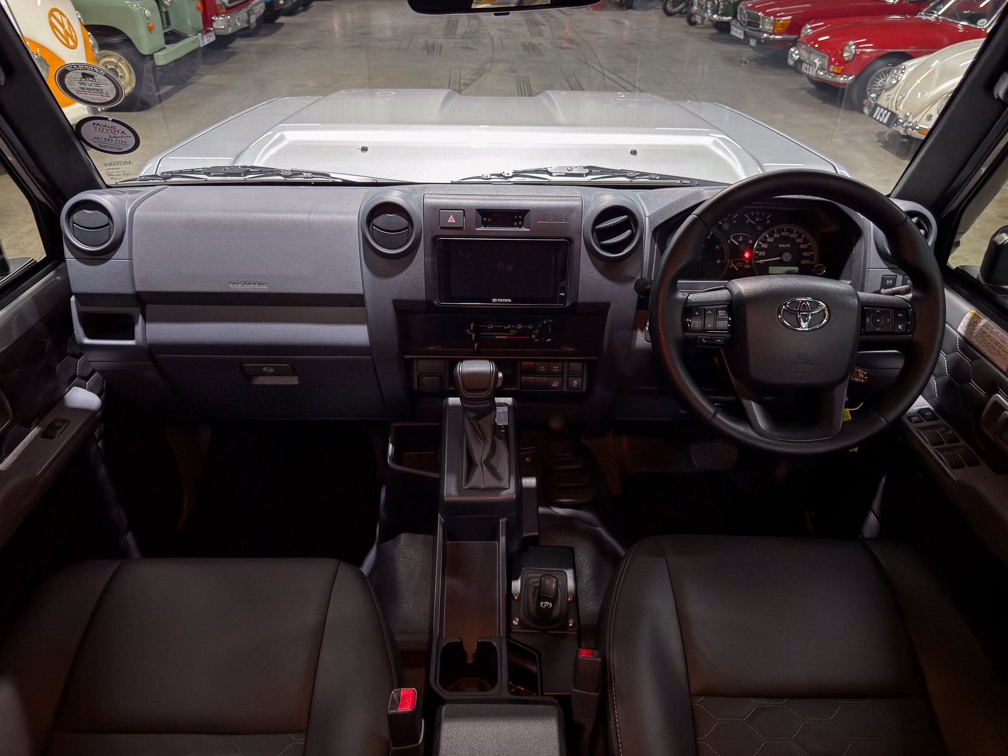 Interior view from the driver's seat showing a right-hand drive dashboard, steering wheel with Toyota logo, center touchscreen, dashboard vents, and gear shifter; showroom with classic cars visible through the windshield.