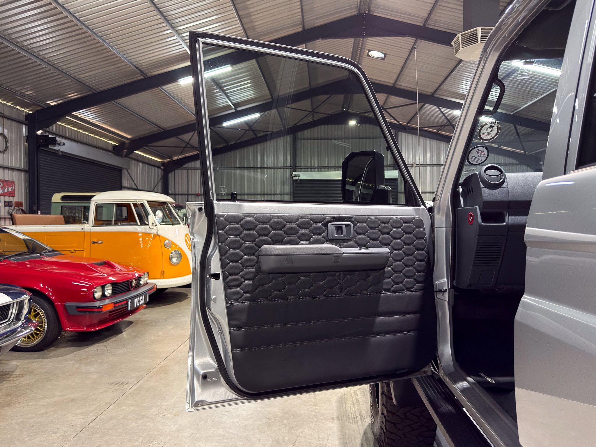 Open SUV door showing a black quilted interior panel in a car showroom with vintage vans in the background, chrome trim visible on the door frame.