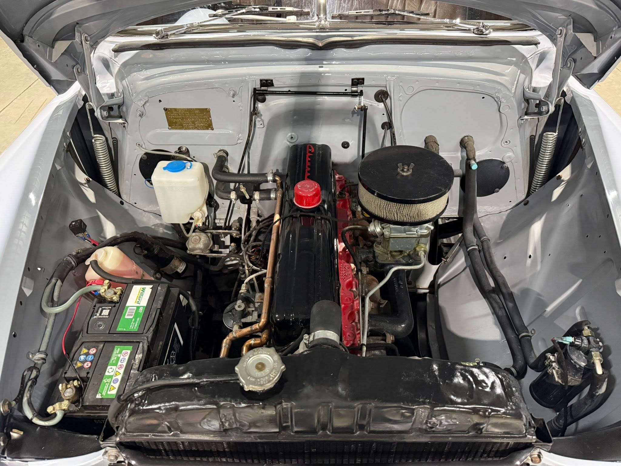 Open car hood showing the engine bay: battery on the left, coolant reservoir, air filter, and various hoses and metal firewall.