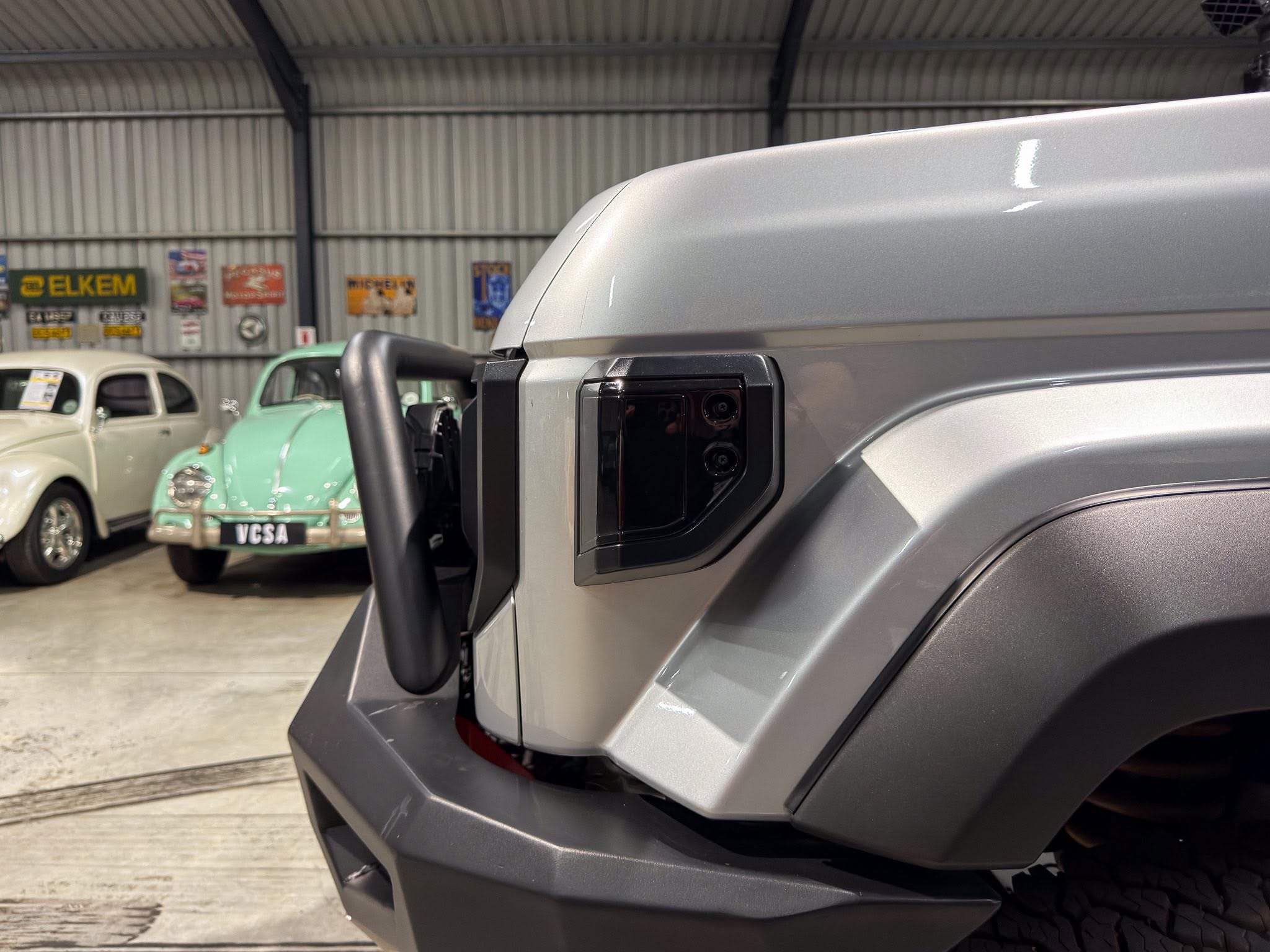 Close-up of a silver vehicle door and side mirror in a showroom, with vintage cars visible in the background.