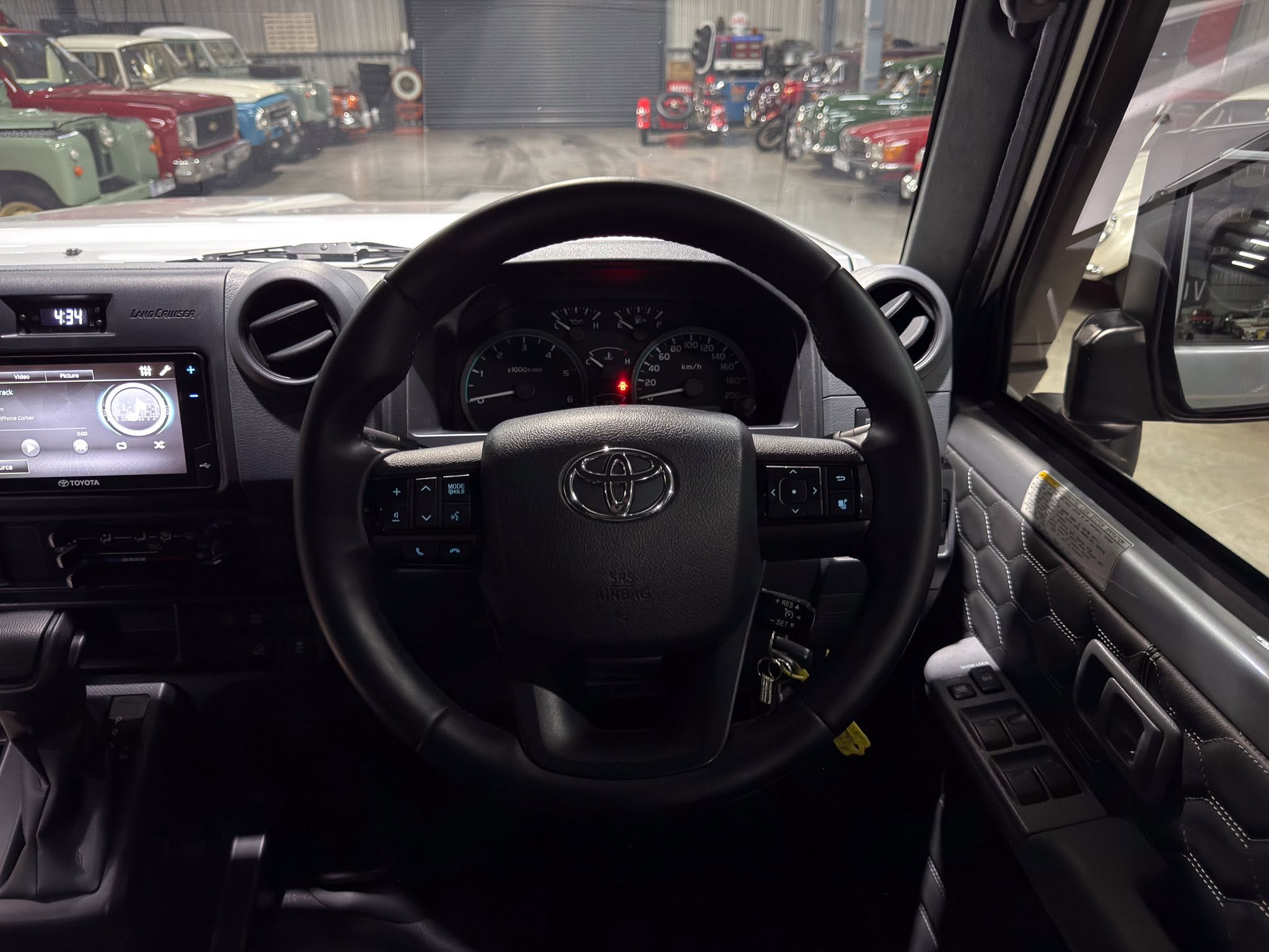 View from the driver's seat of a Toyota, showing the steering wheel and dashboard with a row of vintage cars inside a showroom in the background in a garage setting.