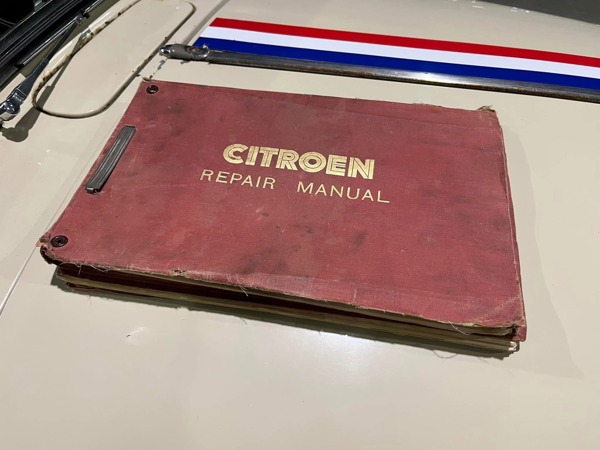 Worn red Citroën repair manual lying on a light-colored car hood with a red-white-blue stripe in the background; visible gold text reads 'CITROEN REPAIR MANUAL'.