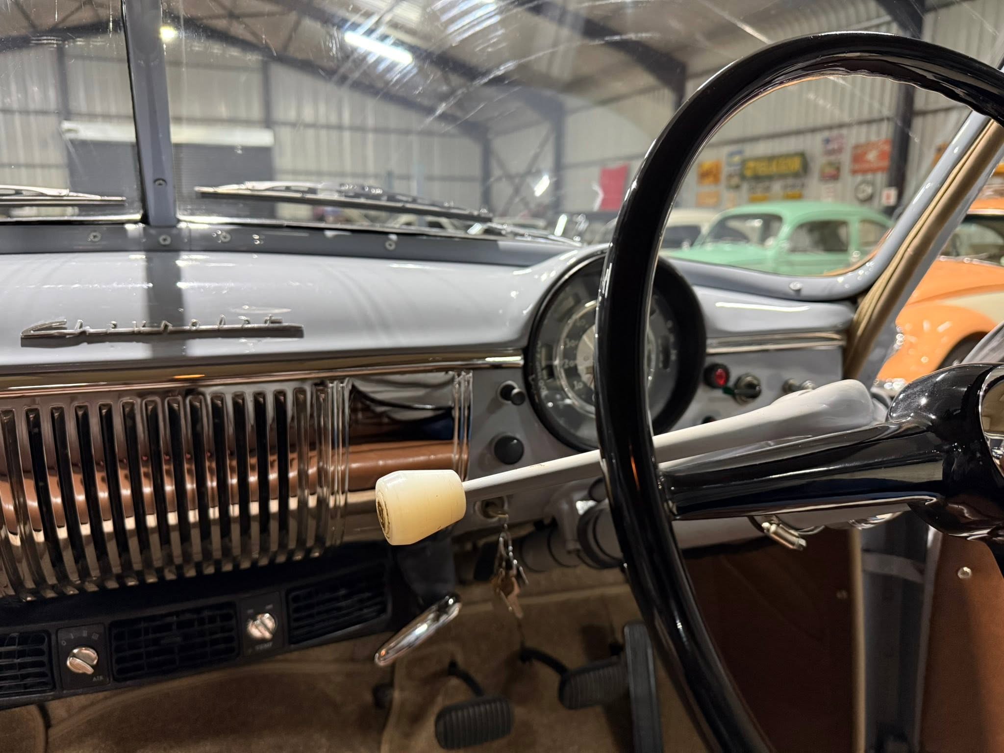 View of a vintage car interior, showing a chrome-grilled dashboard, gray panel, and beige shift knob, with other classic cars in a showroom visible through the windshield.
