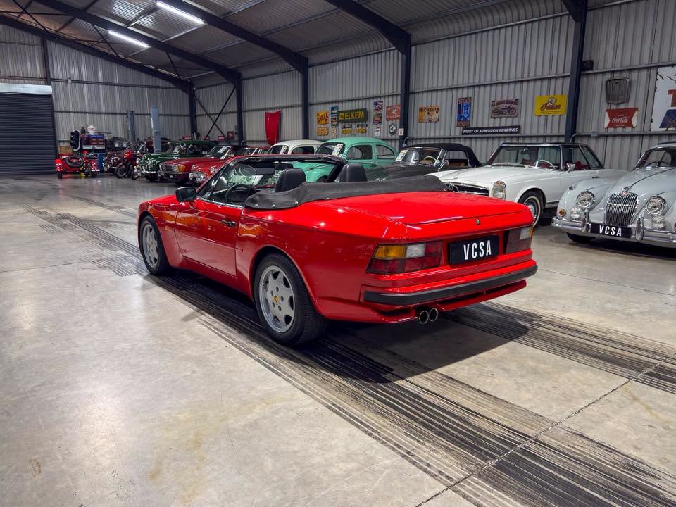 Red convertible sports car with its black soft top folded down in a vintage car showroom surrounded by classic cars.