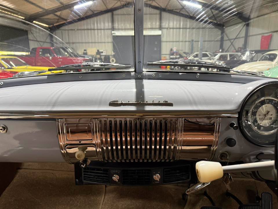 Inside a classic car cockpit with chrome grille detail on the dashboard and a large round speedometer on the right, photographed in a showroom.