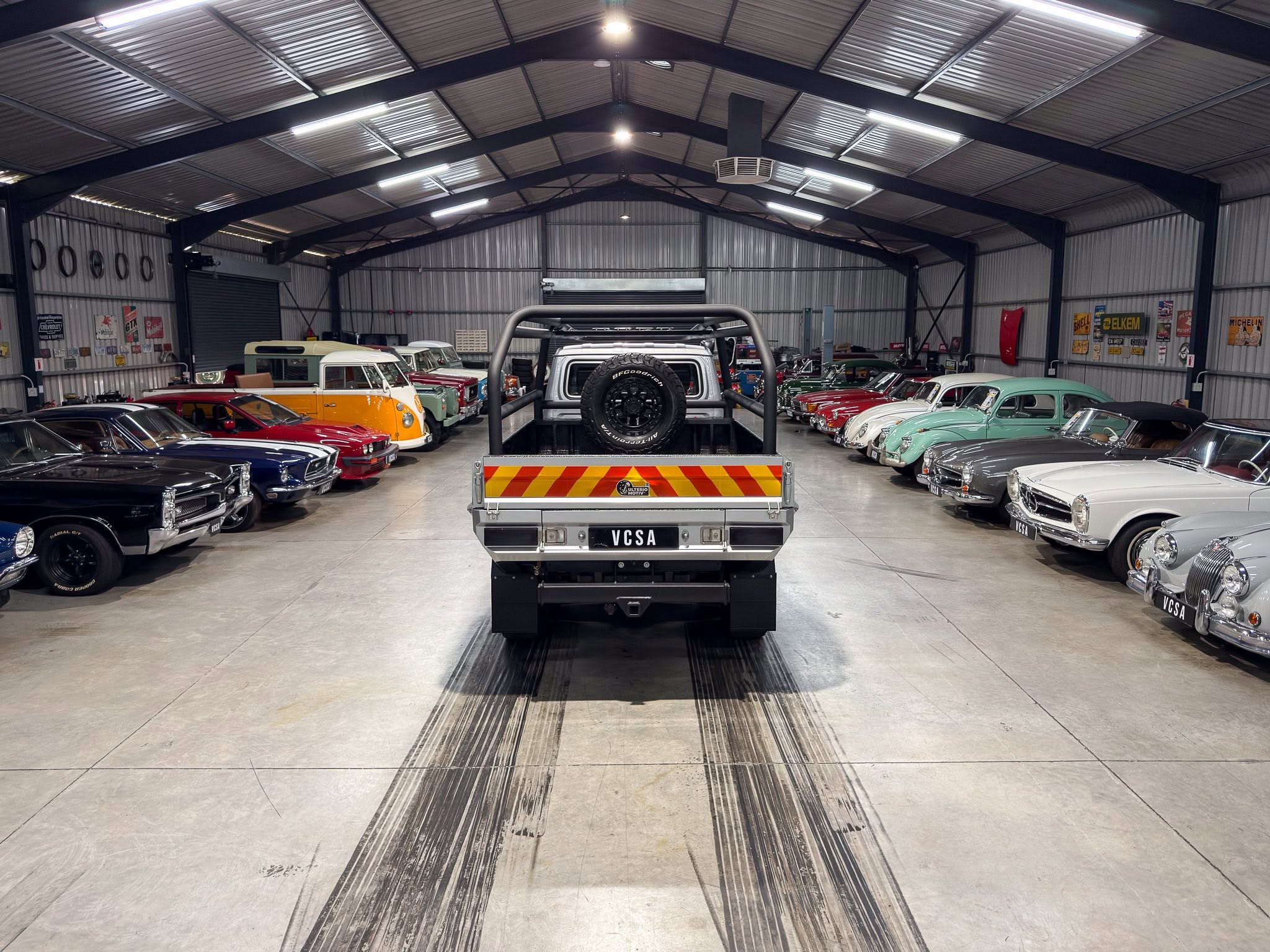 Indoor showroom of vintage cars, with a silver pickup centered and spare tire on back, rows of classic vehicles on both sides under a metal roof.
