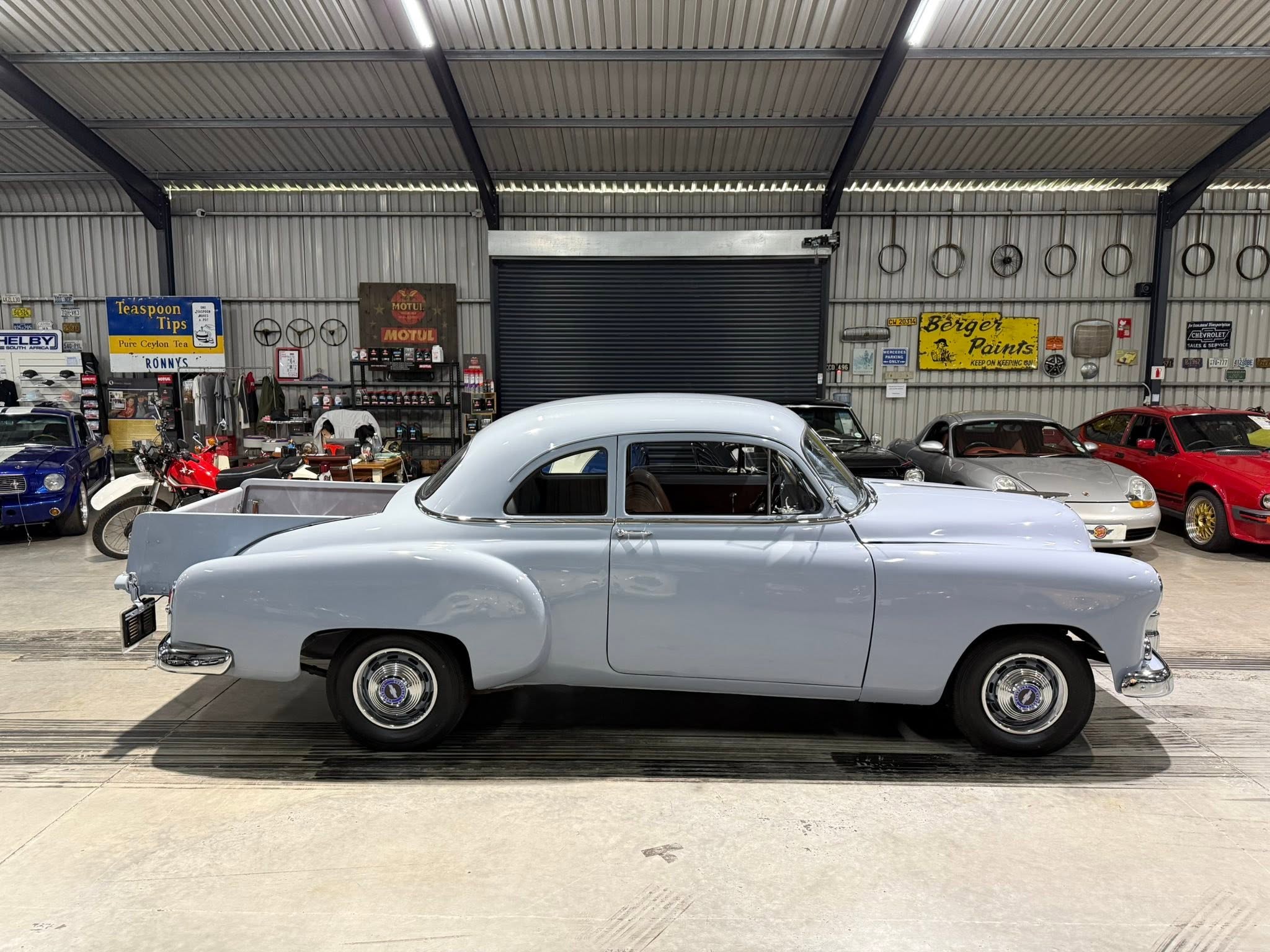 Inside a vintage car workshop with several classic cars parked and retro signs on metal walls, including a blue bike and a pale blue coupe in the foreground.