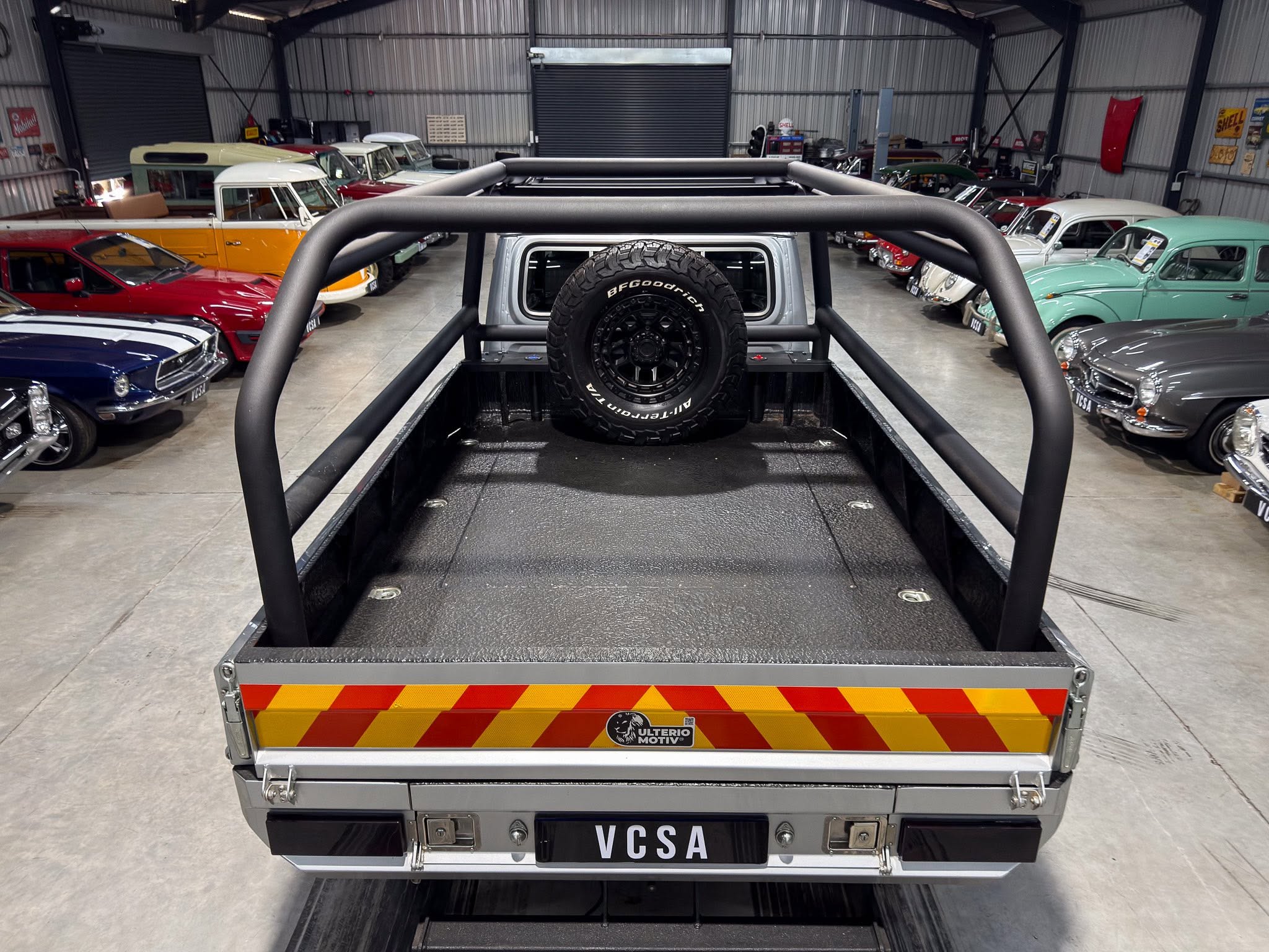 Rear view of a utility trailer with a mounted spare tire inside a showroom filled with vintage cars on both sides.