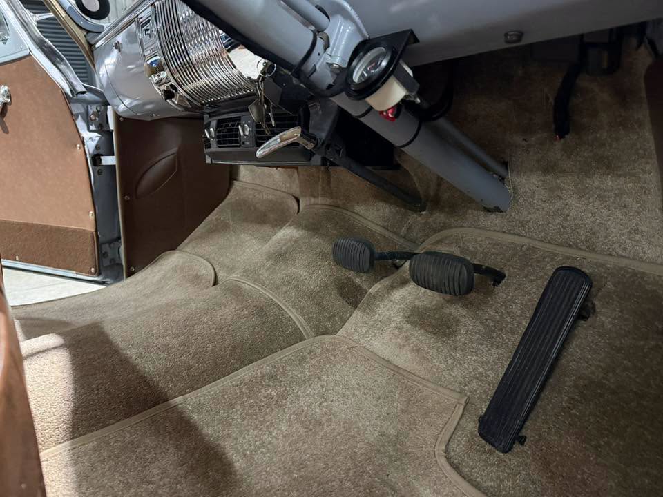 Driver's footwell in a vintage car showing three pedals: clutch, brake, and accelerator on tan carpet with chrome dash details nearby.