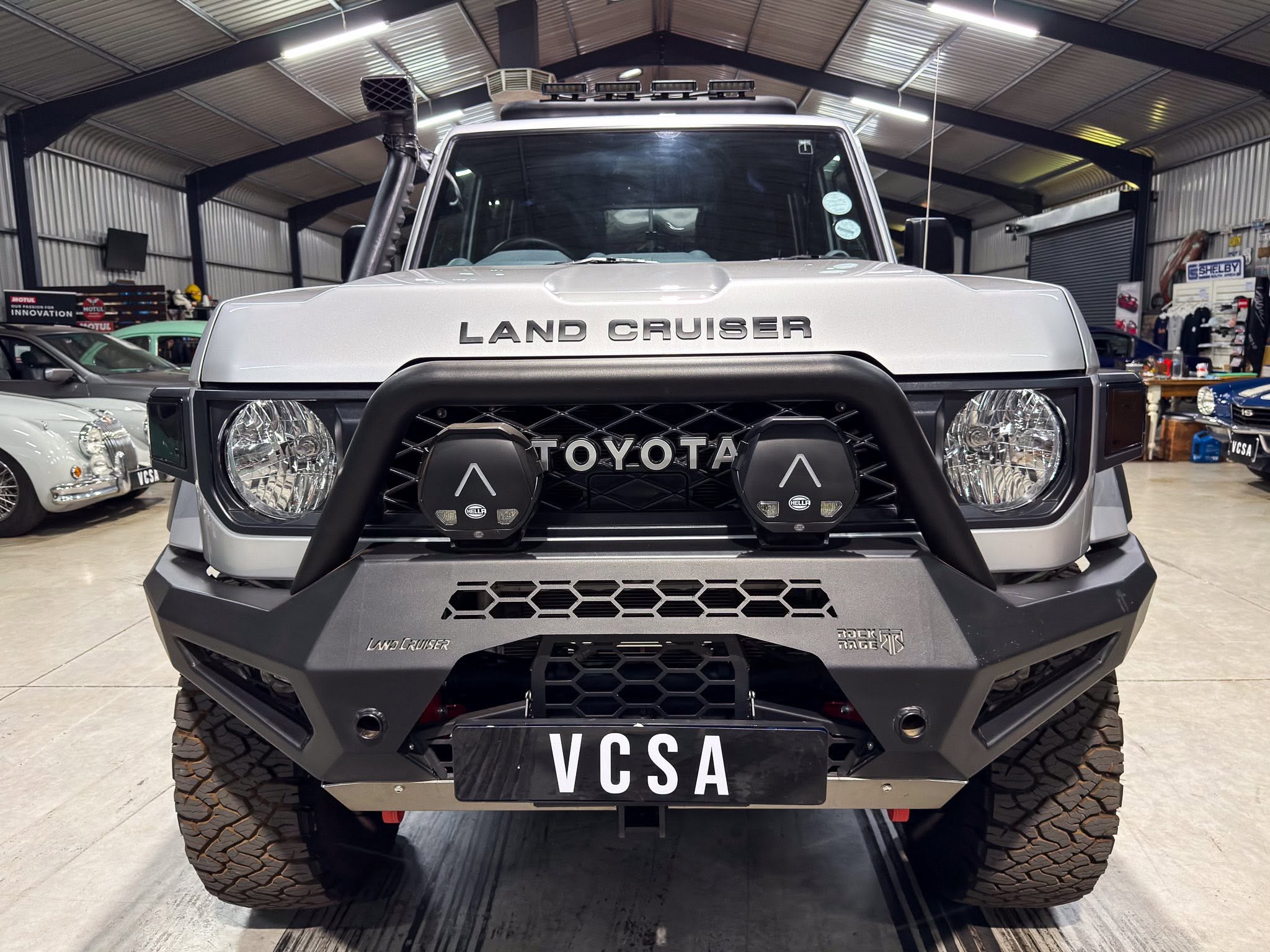 Front view of a Toyota Land Cruiser in a showroom, featuring an aftermarket heavy-duty grille guard and two round off-road lights; license plate reads VC SA.