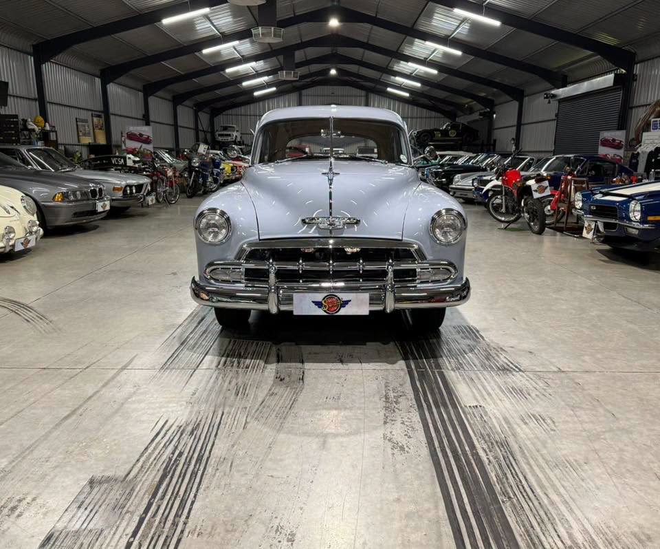 Front view of a vintage silver Chevrolet parked inside a large showroom filled with classic cars on both sides.