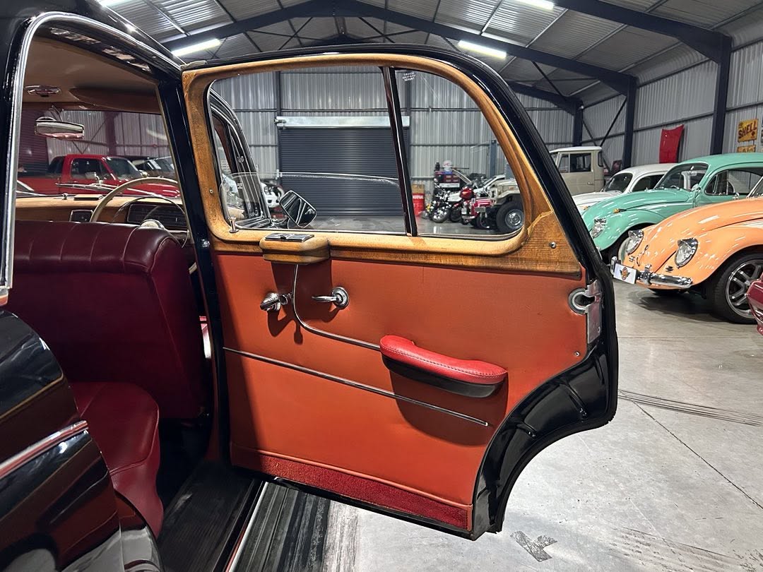 Open rear door of a vintage car showing red leather interior and wood trim, inside a showroom with other classic cars visible in the background.
