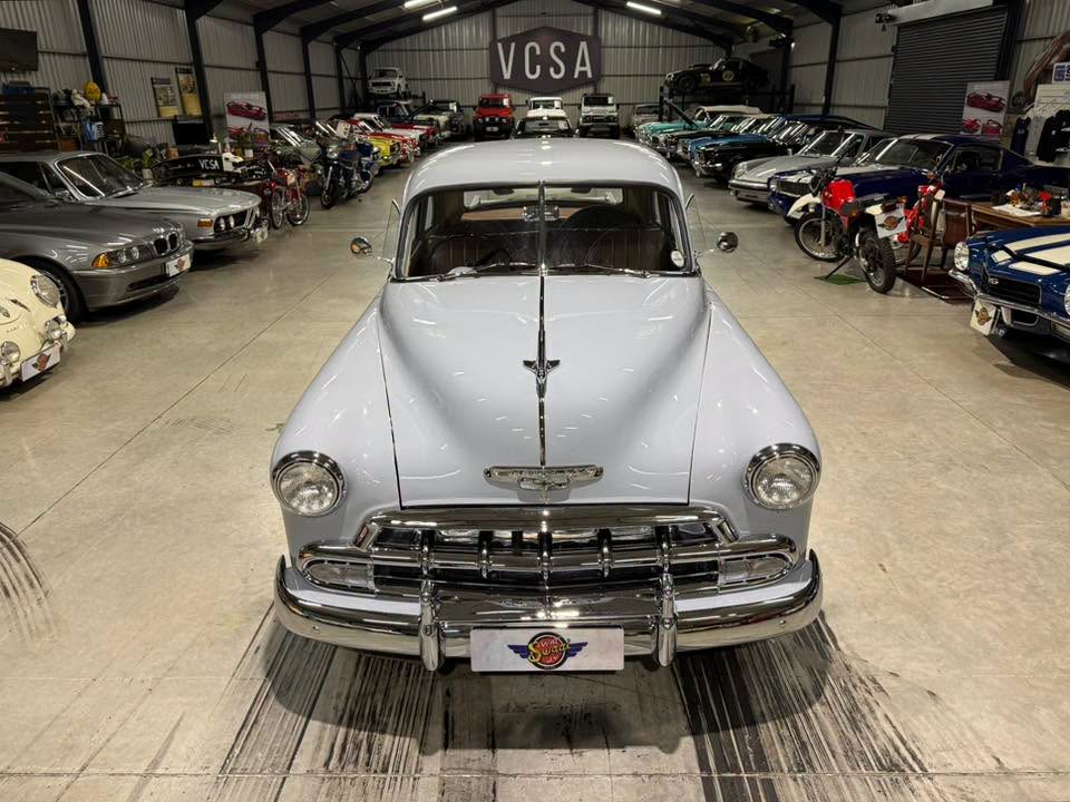 Front view of a pale blue vintage car with chrome bumper, parked in a large showroom filled with classic cars.