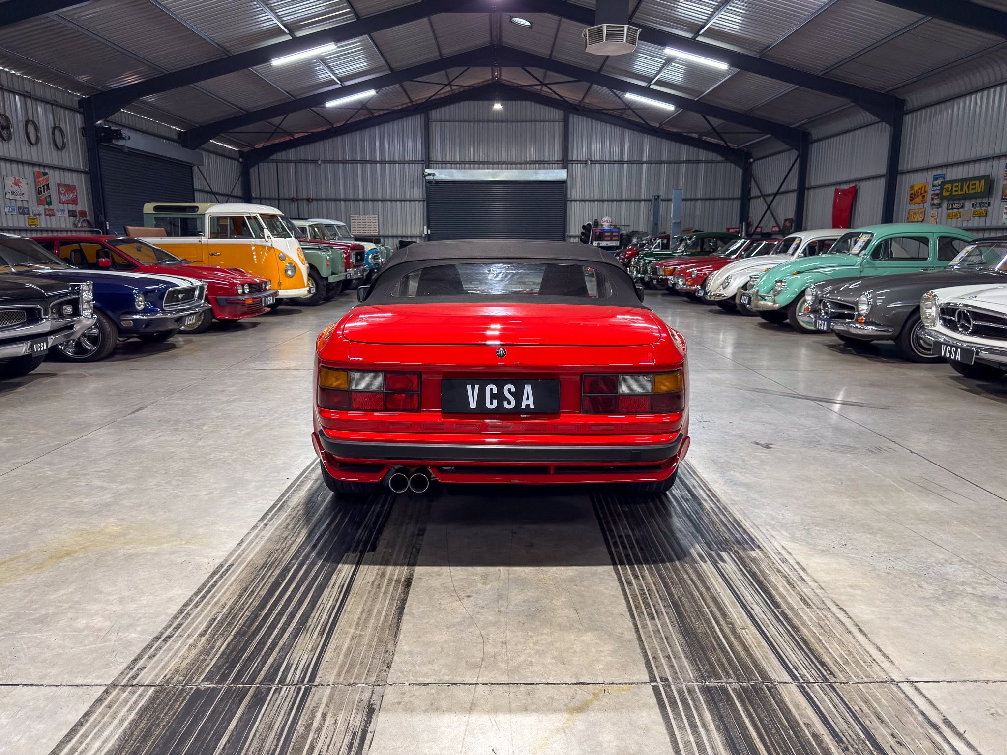 Red vintage convertible viewed from behind in a large car showroom with classic cars on both sides under a metal warehouse roof.