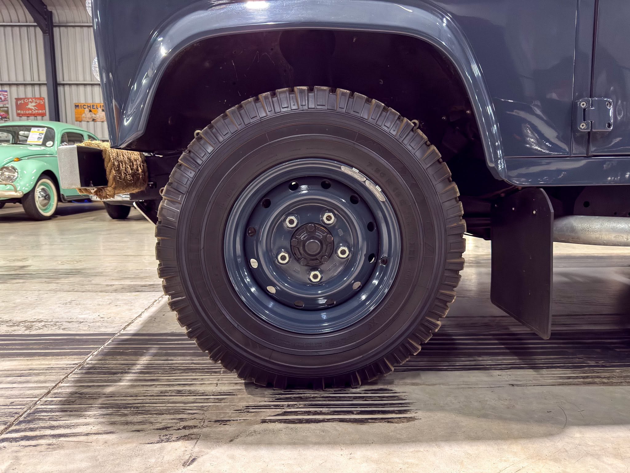 Close-up of a rugged off-road tire on a dark blue vehicle in a workshop; a vintage green car is visible in the background.