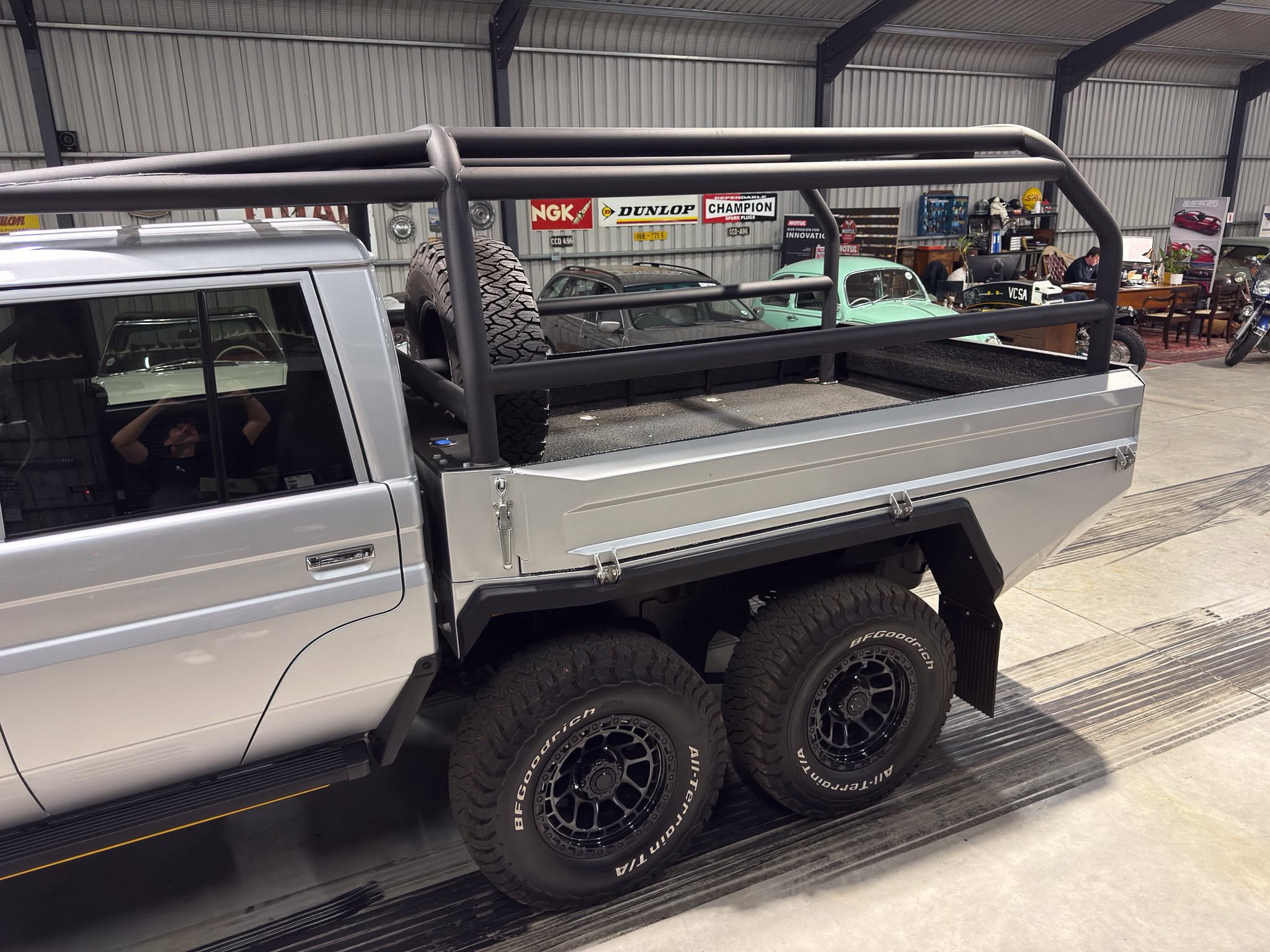 Silver pickup truck with a large, black tubular bed rack and spare tire mounted in the truck bed inside a workshop displaying vintage cars and banners on the wall