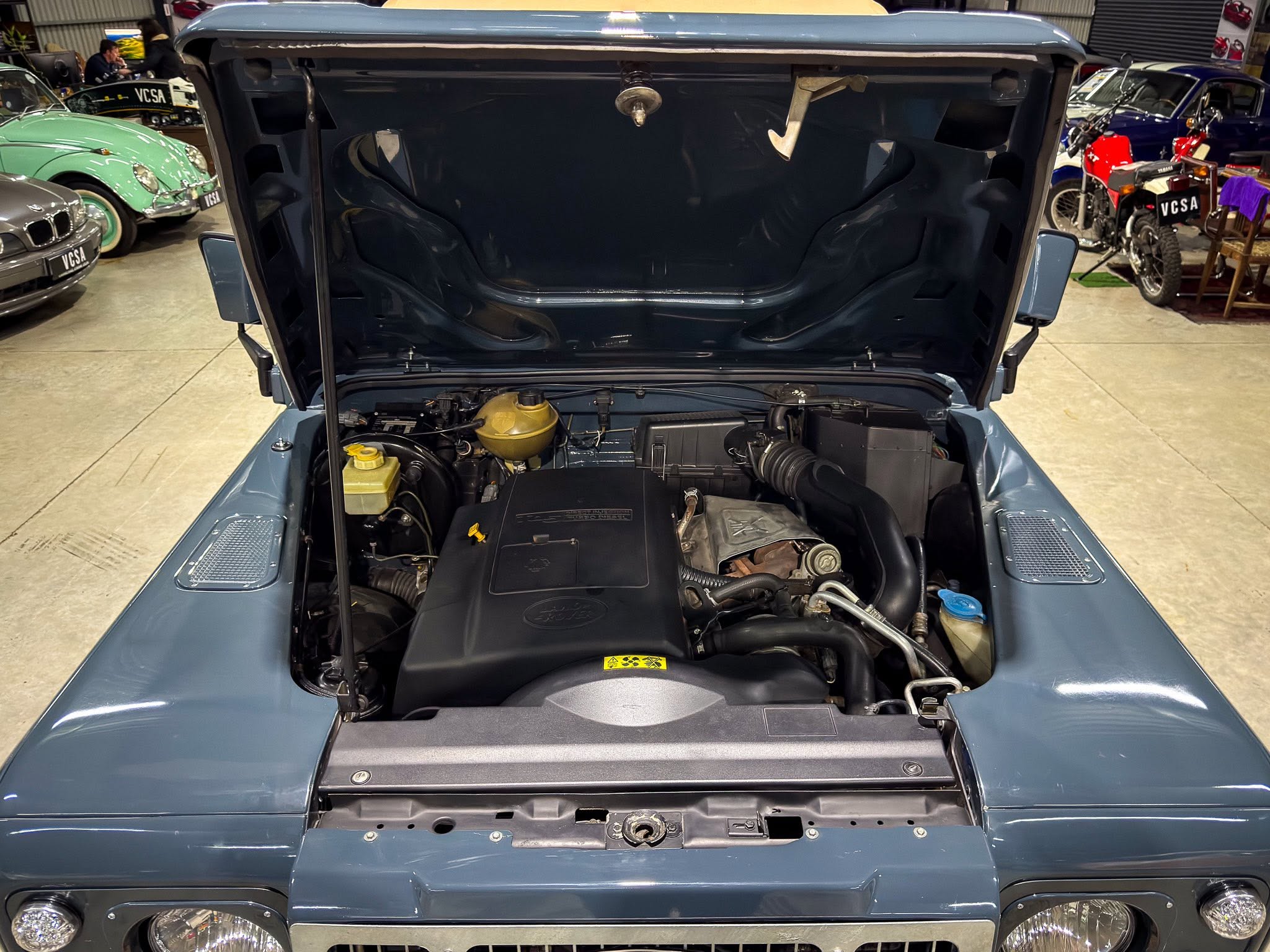 Blue classic SUV with hood open in a showroom, exposing the engine bay and components, with other vintage cars visible in the background