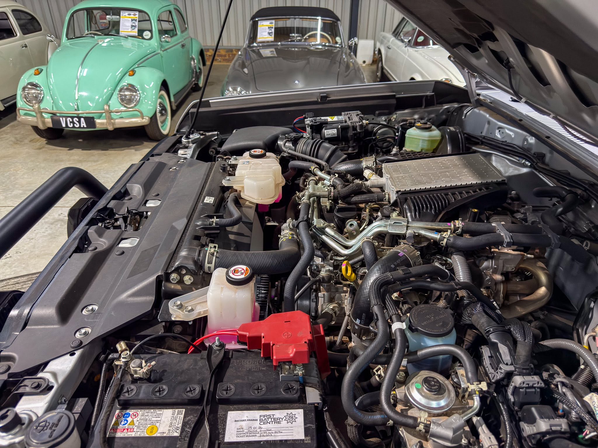 Open hood of a modern car in a garage or showroom, exposing the engine and components; in the background are vintage cars, including a mint-green Volkswagen Beetle and other classics.