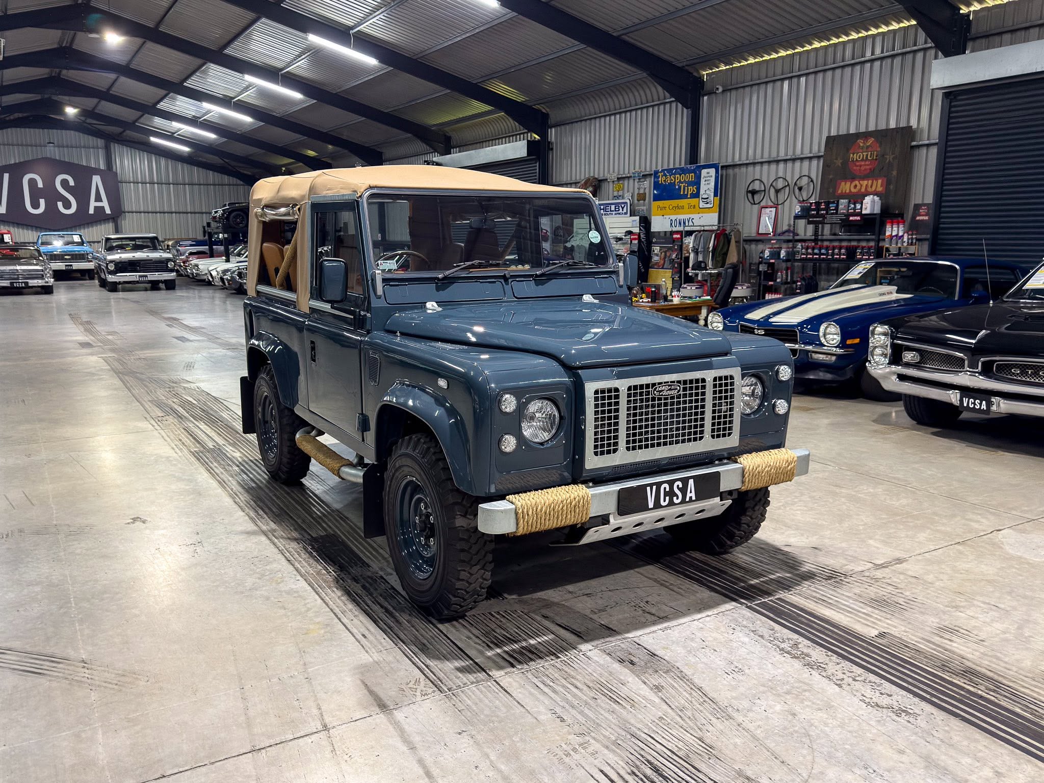 Indoor car showroom featuring a blue Land Rover Defender in the foreground with a tan soft top and other classic cars in the background (VCSA plates).