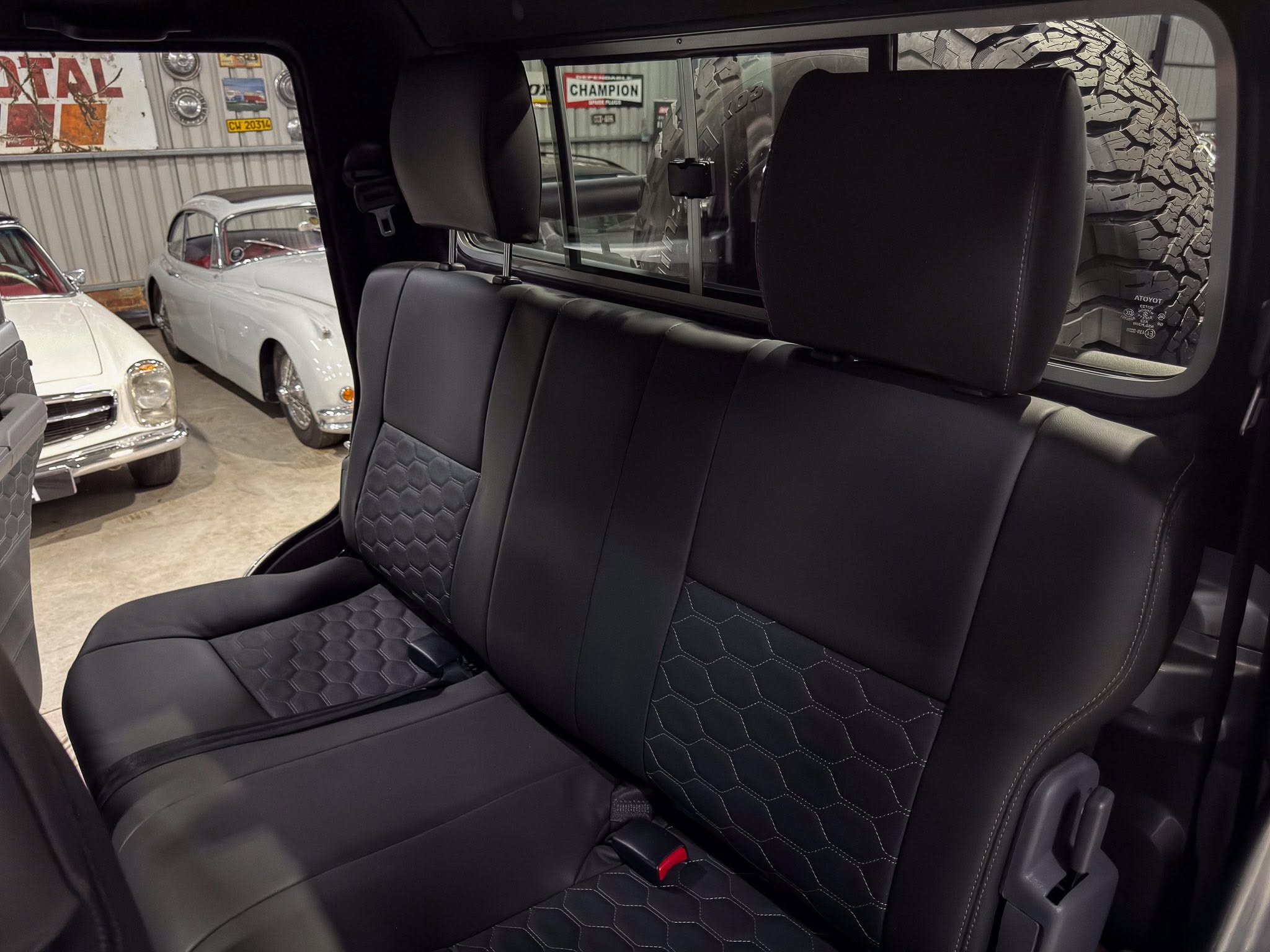 Back seat of a black leather car with diamond-stitched upholstery and headrests, viewed from the rear passenger area with classic cars outside the window.