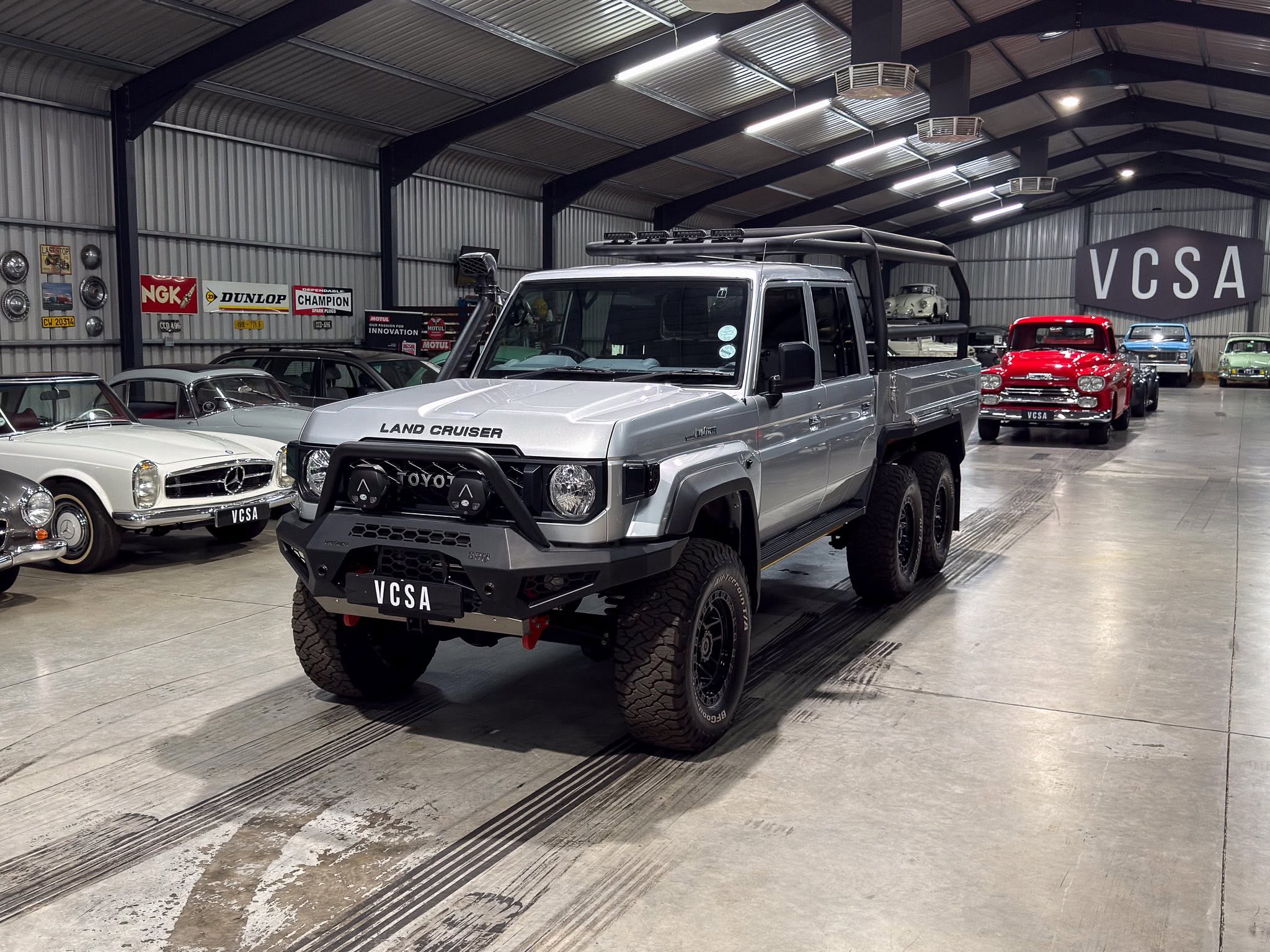 Indoor car showroom with a silver Toyota Land Cruiser in the foreground and vintage cars along the walls under a metal ceiling, plus a large VCSA sign in the background.